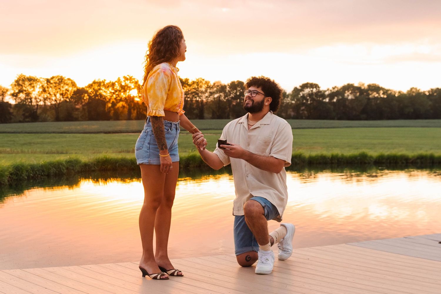 Man proposing on one knee to woman on a wooden dock during a vibrant sunset in Elverson, PA.