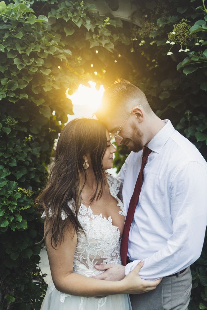 Bride and groom share a romantic moment surrounded by lush greenery with golden sunlight glowing behind them.