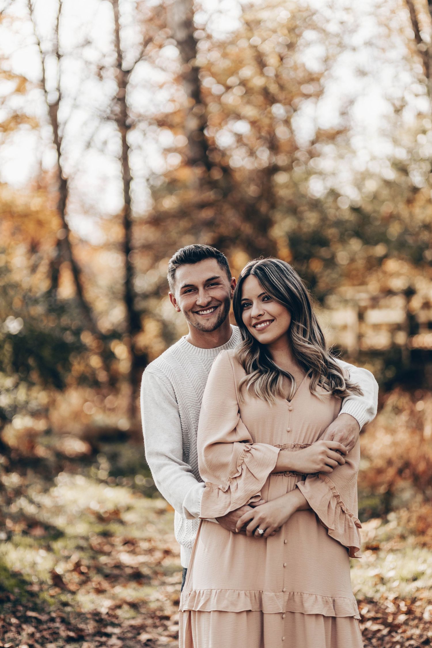Smiling couple embracing in autumn forest, man in cream sweater hugging woman in pink dress.