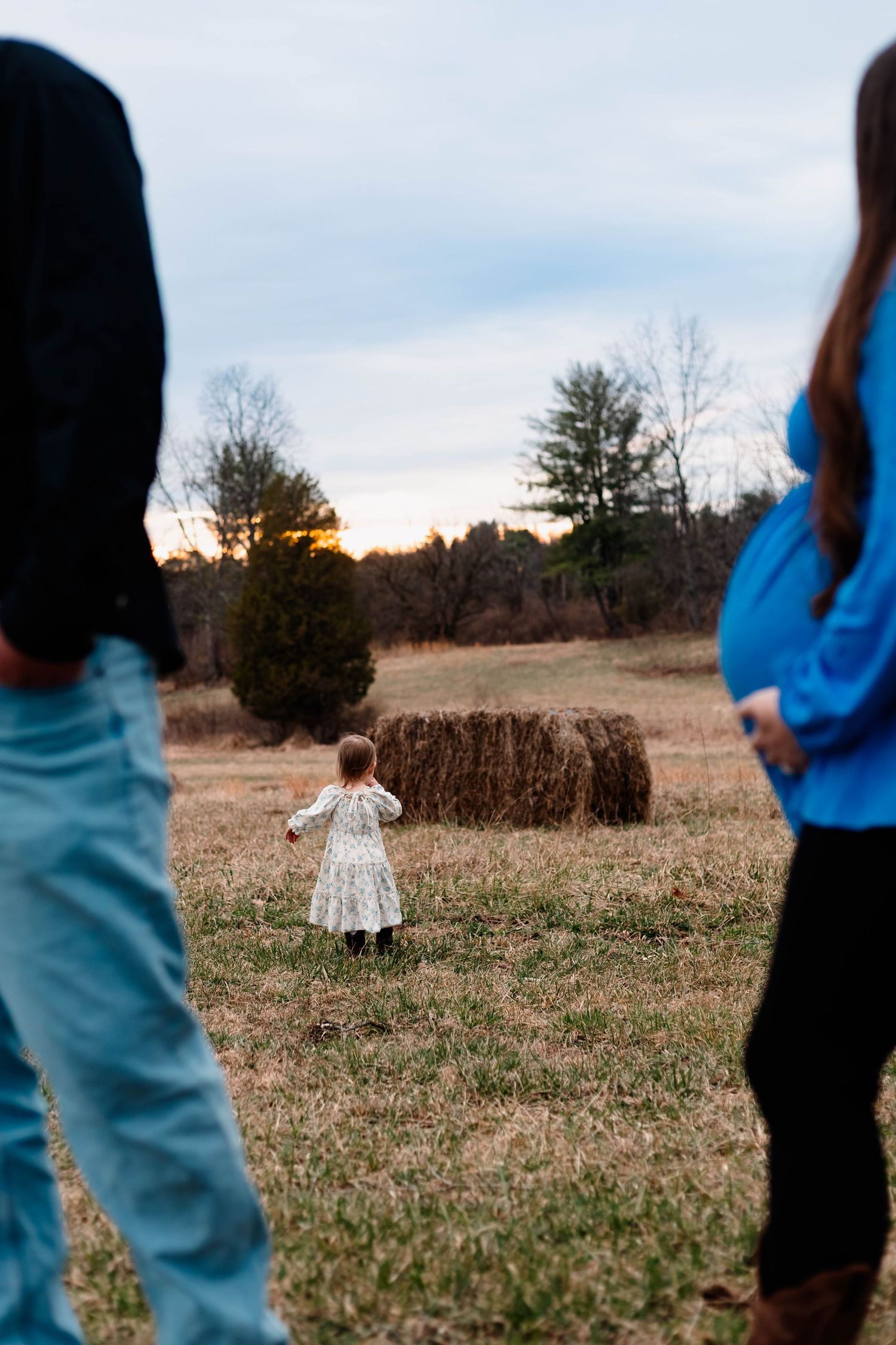 Young child in dress walking toward hay bale at sunset between expecting parents outdoors at Daniel Boone Homestead.