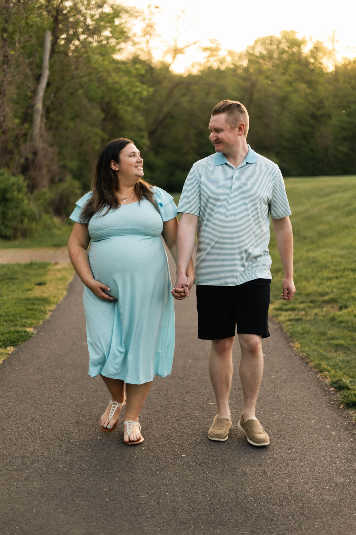 Smiling expecting couple holding hands and walking together on a park path at golden hour in Philadelphia, PA.