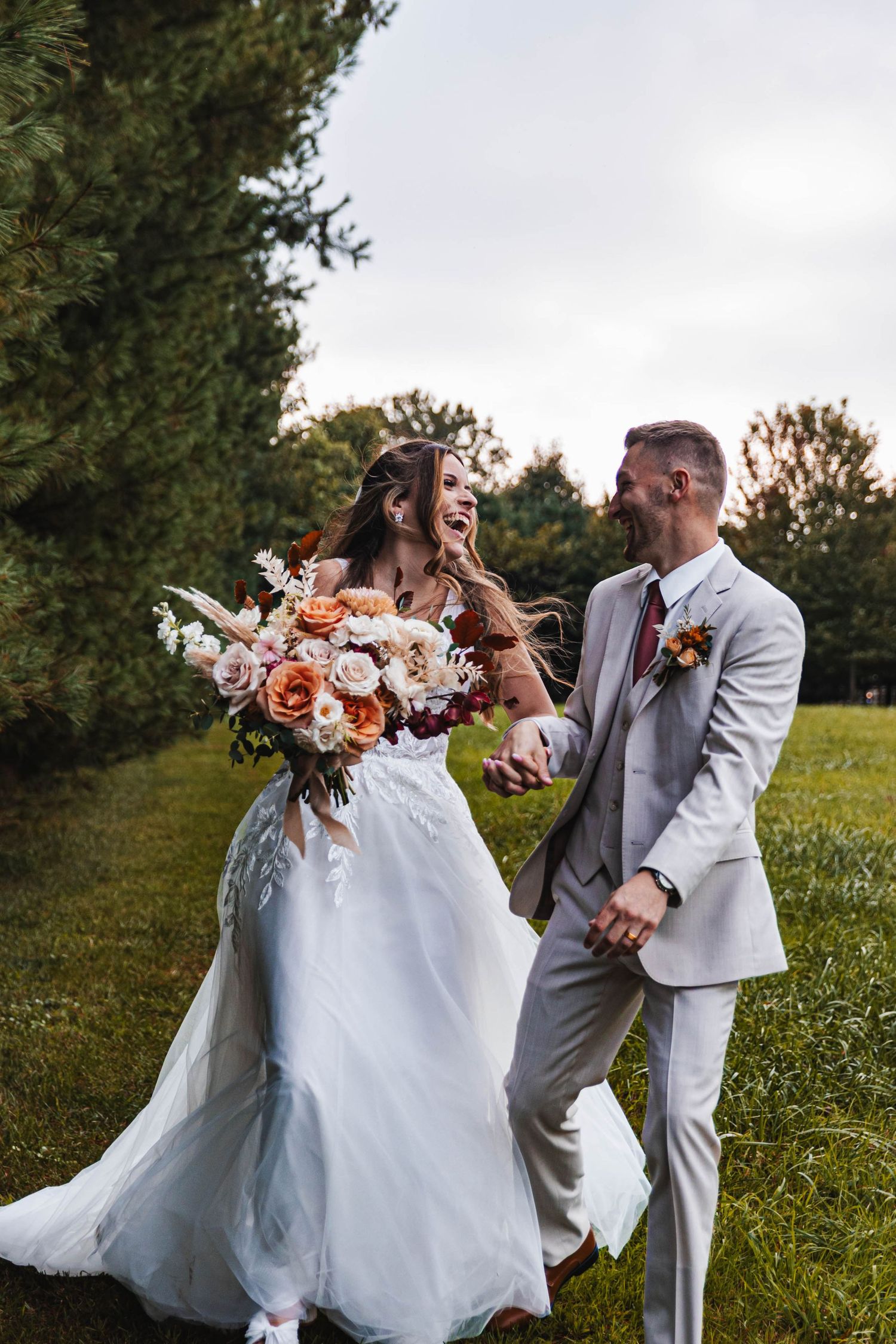 Laughing bride in lace gown holds colorful bouquet while running with groom in gray suit outdoors.