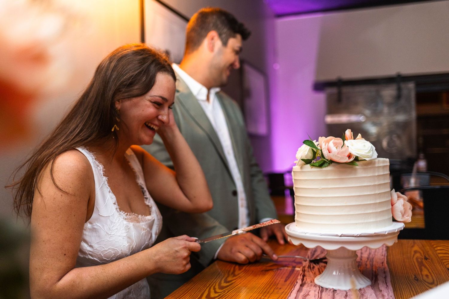Laughing bride cutting white tiered wedding cake decorated with pink flowers at reception.