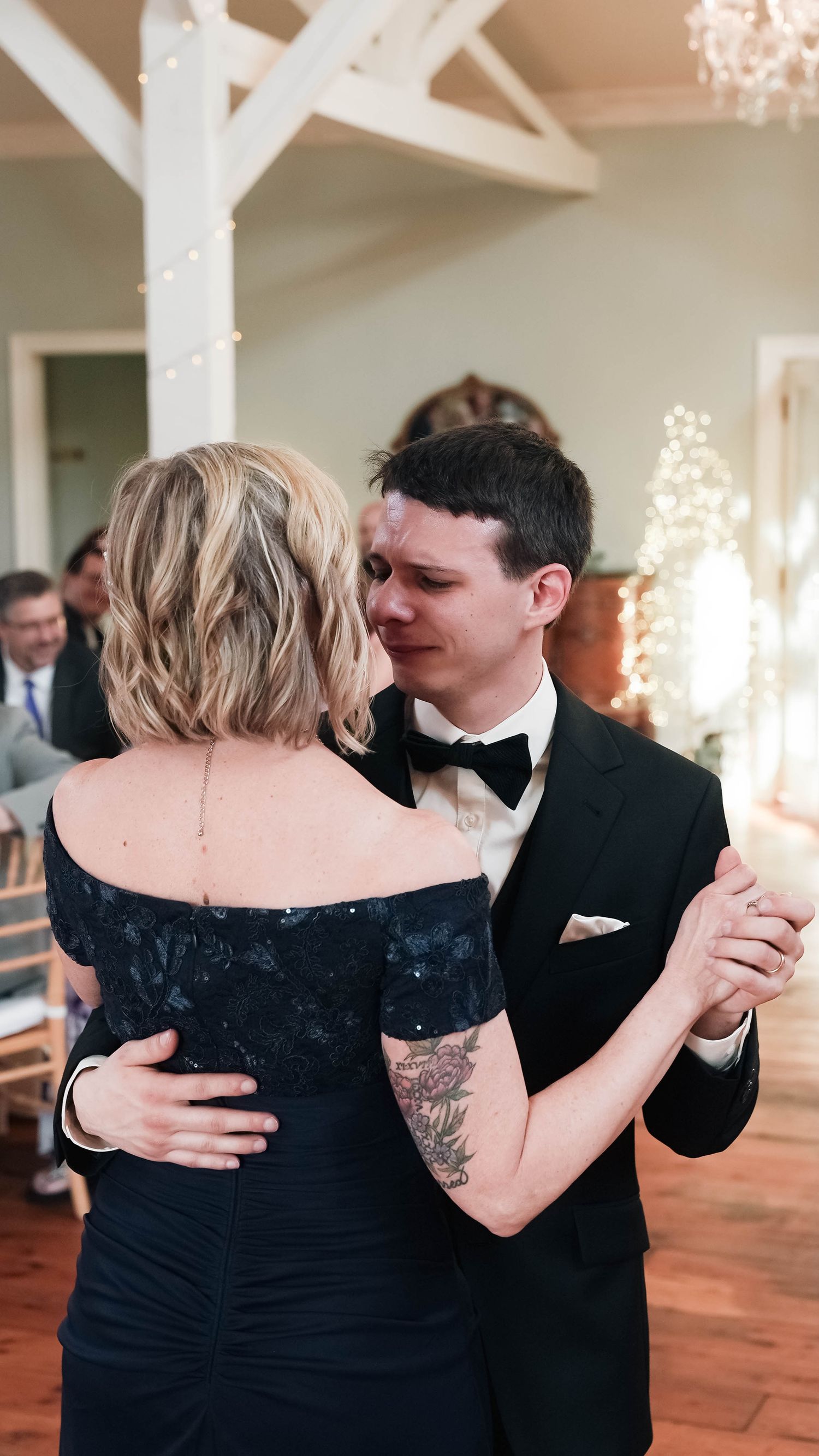 Emotional groom in black tuxedo shares mother-son dance at elegant wedding reception with string lights.