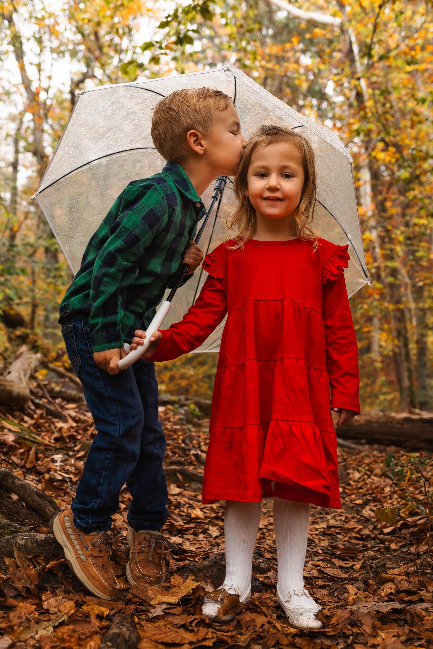 A young boy kisses his sister on the cheek as they share an umbrella among fall leaves in a forest in Pottstown, PA.