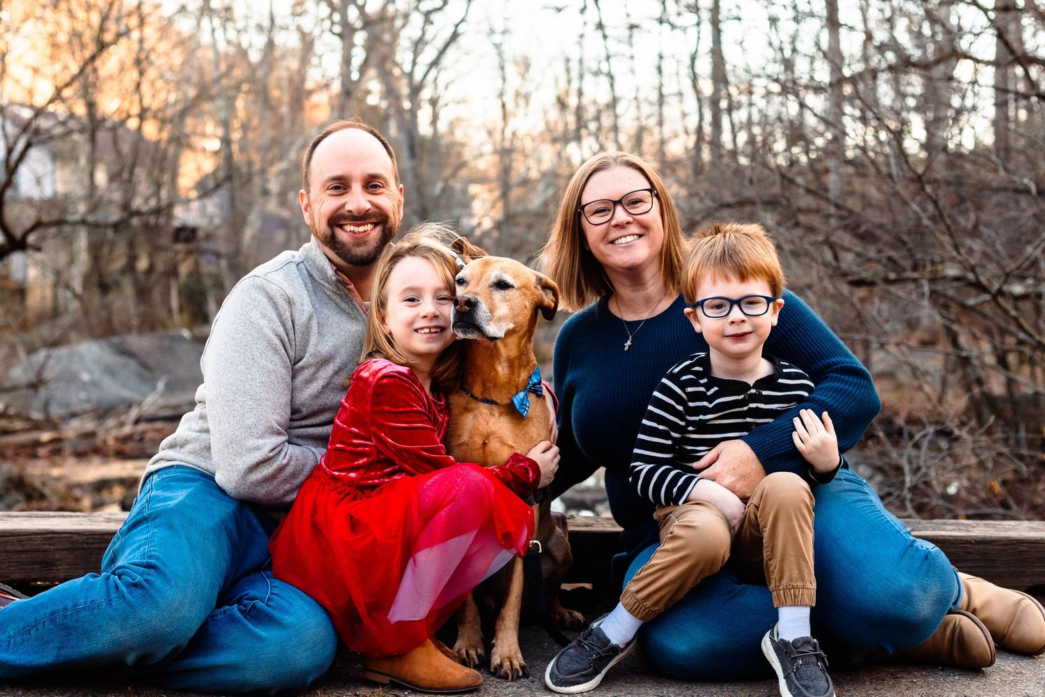 A smiling family of four with a golden retriever wearing a bow tie poses outdoors in autumn woods in Pottstown, PA.
