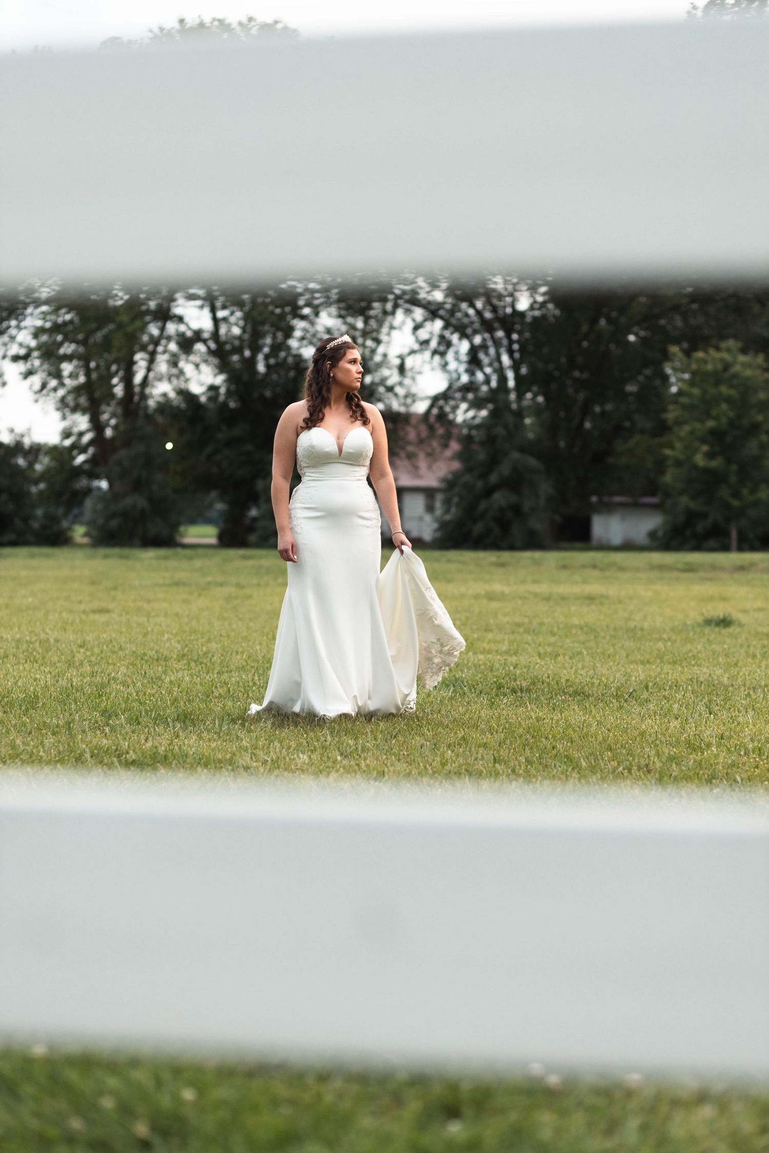 Elegant bride in a fitted white gown stands alone in an open green field framed by white fence rails.