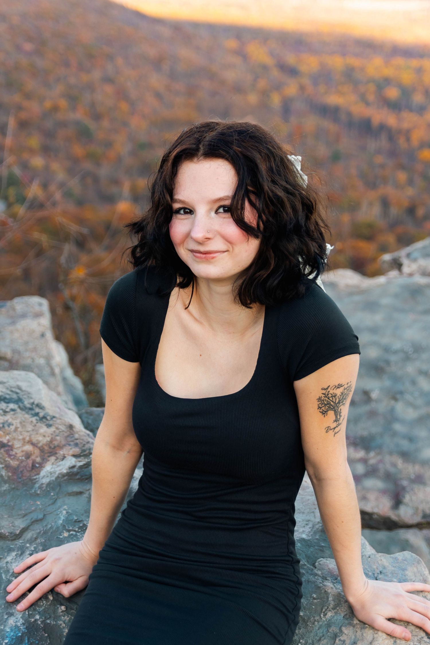 Young brunette woman in black dress sitting on rocks with autumn mountain landscape behind her.