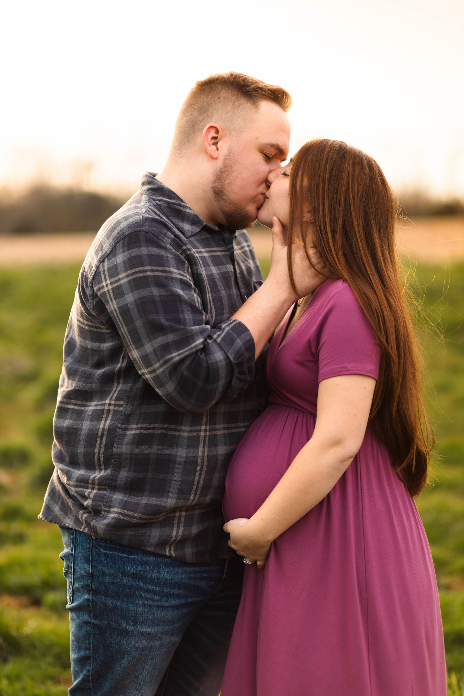 Couple sharing a kiss outdoors at sunset, pregnant woman in purple dress holding baby bump.