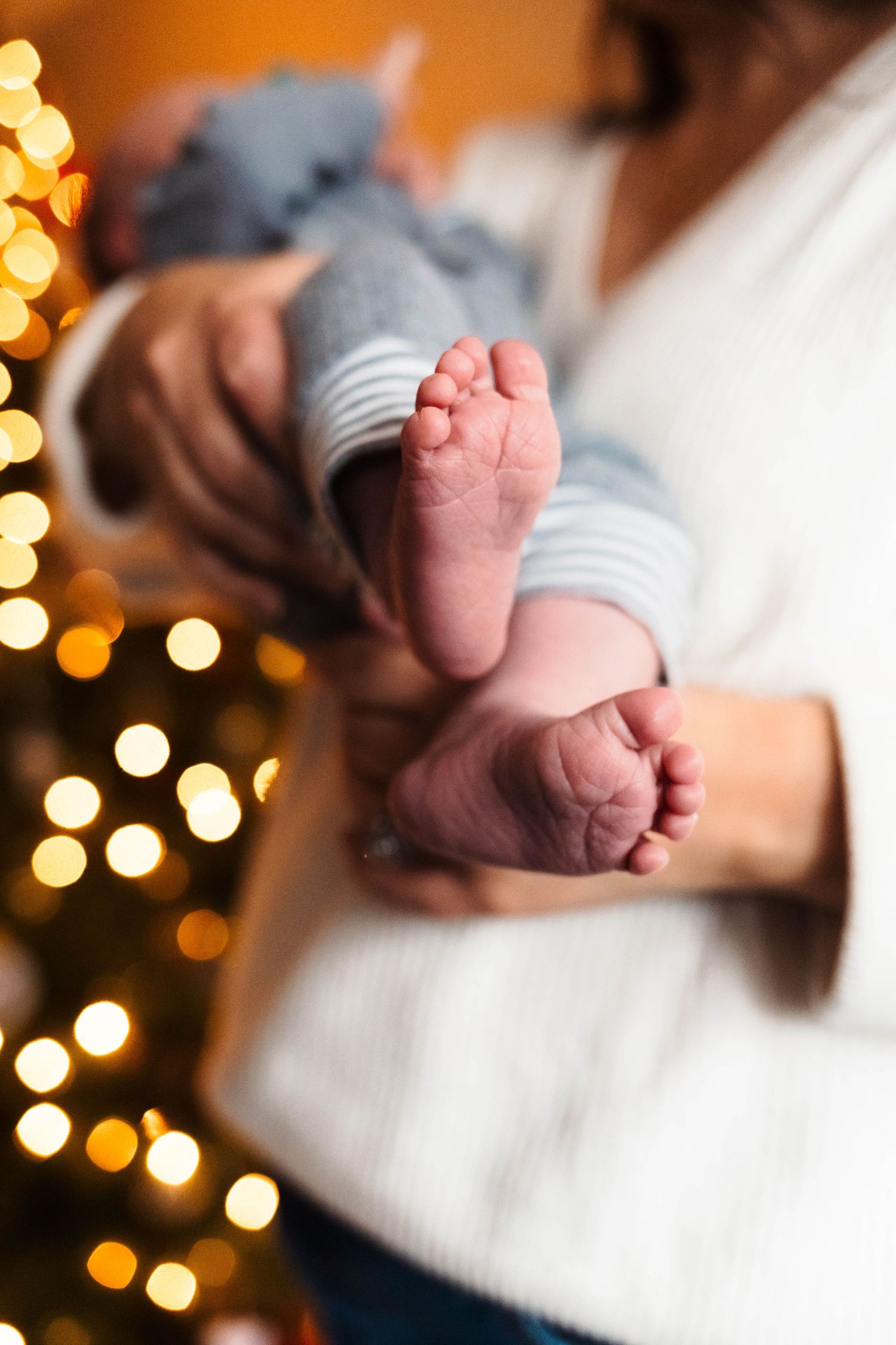 Close-up of newborn baby feet held by mother with bokeh Christmas tree lights in background.