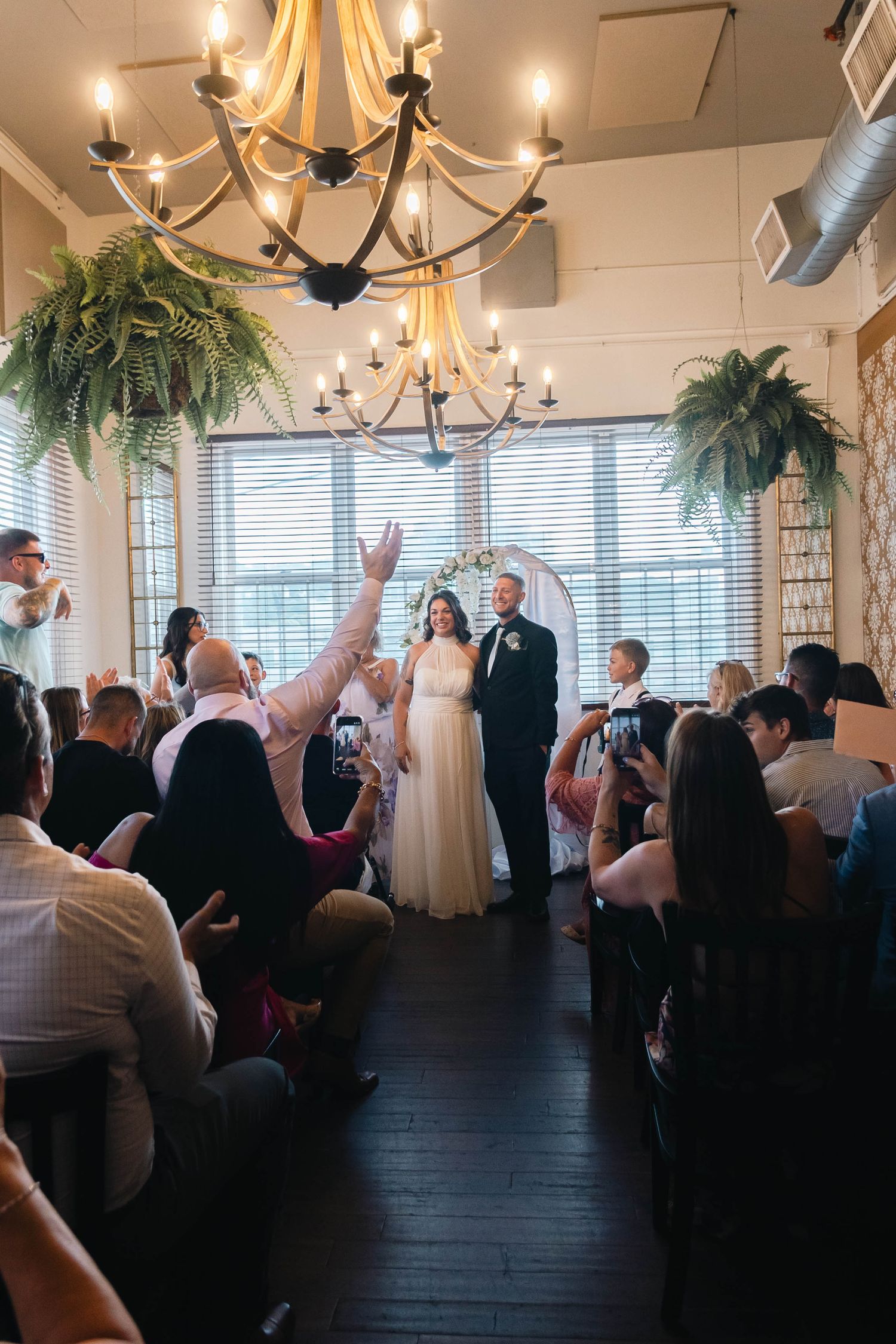 Bride and groom celebrated by guests during indoor wedding ceremony under elegant chandelier with fern decor.