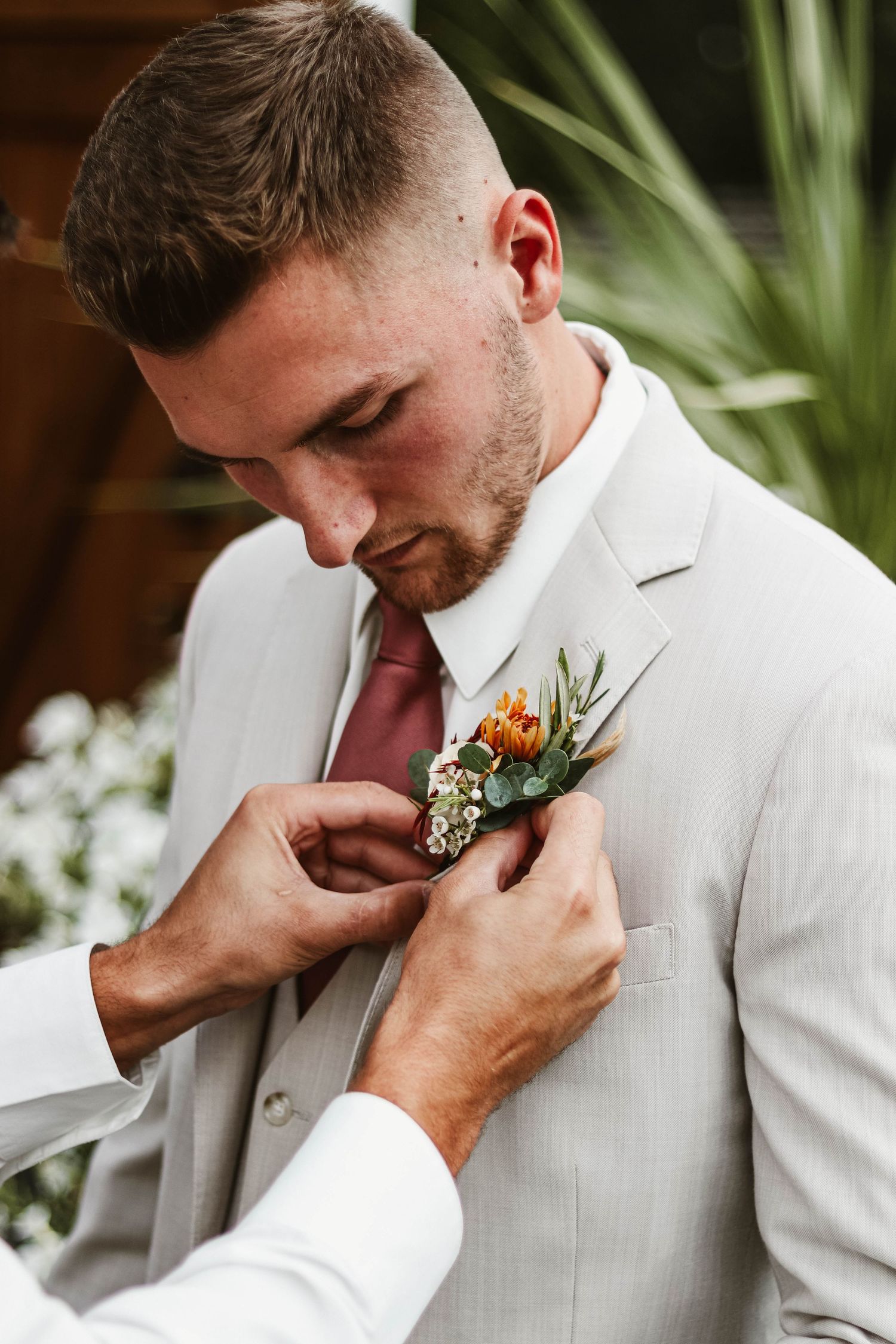 Groomsman pinning orange boutonniere on groom in light gray suit with burgundy tie.