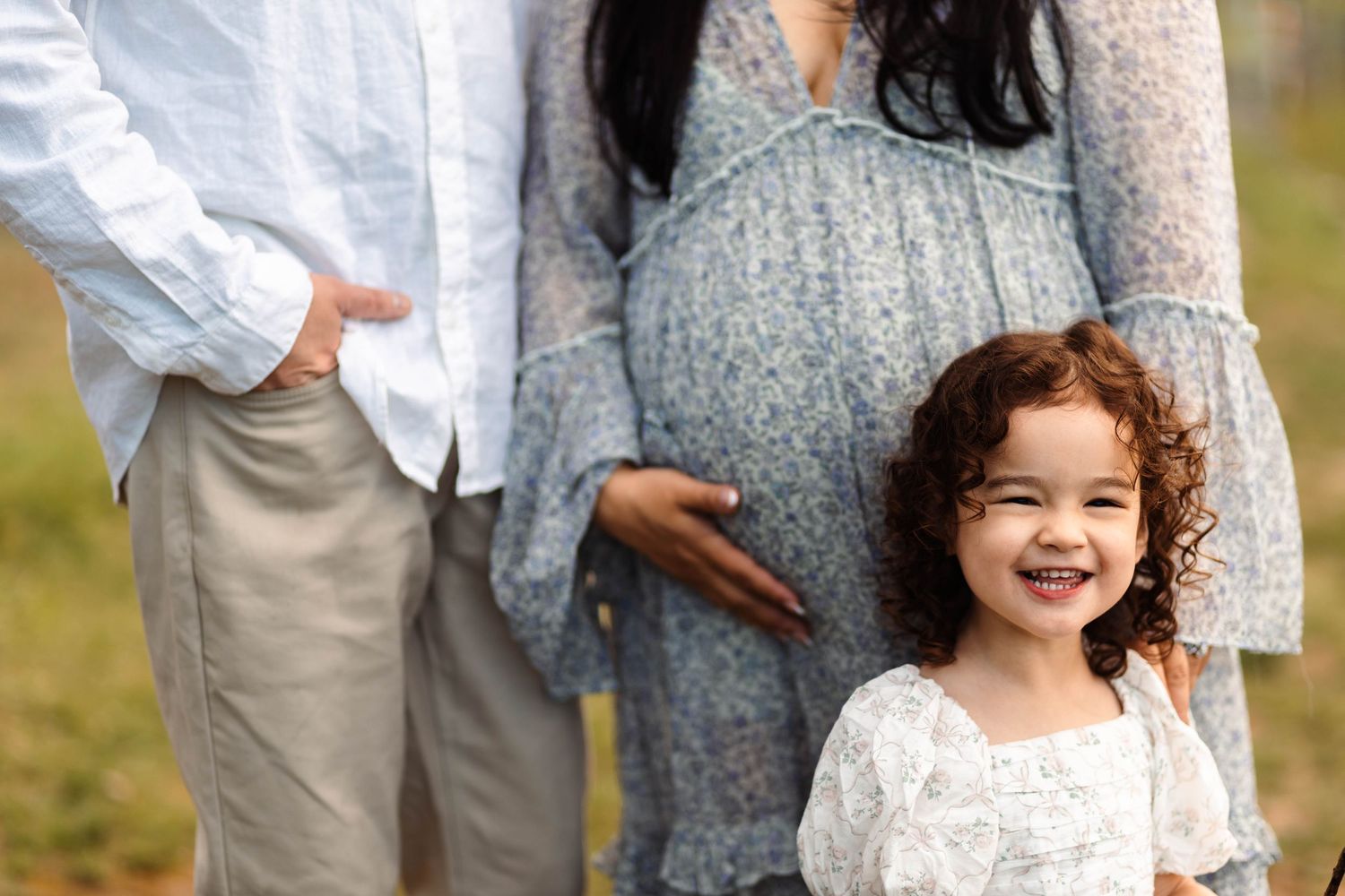 Smiling curly-haired toddler girl stands in front of pregnant mother in blue floral dress outdoors at Green Lane Park.