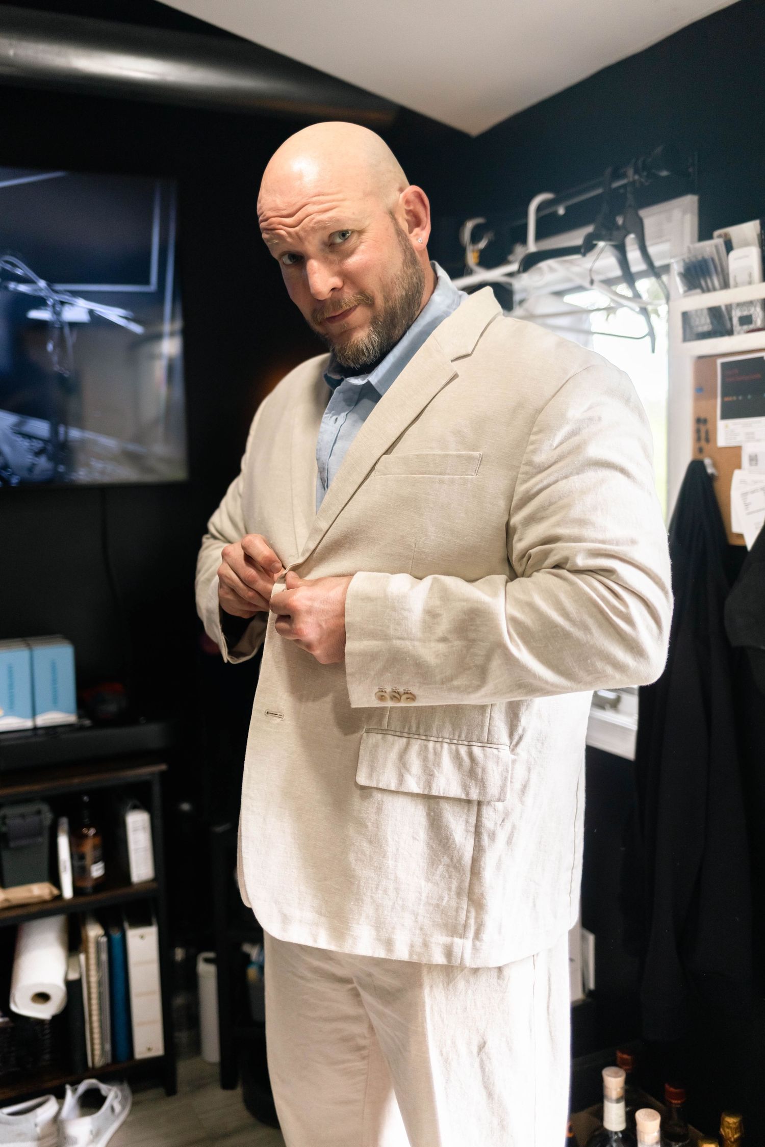 Bald groom in beige linen suit buttoning jacket in dressing room before wedding ceremony.
