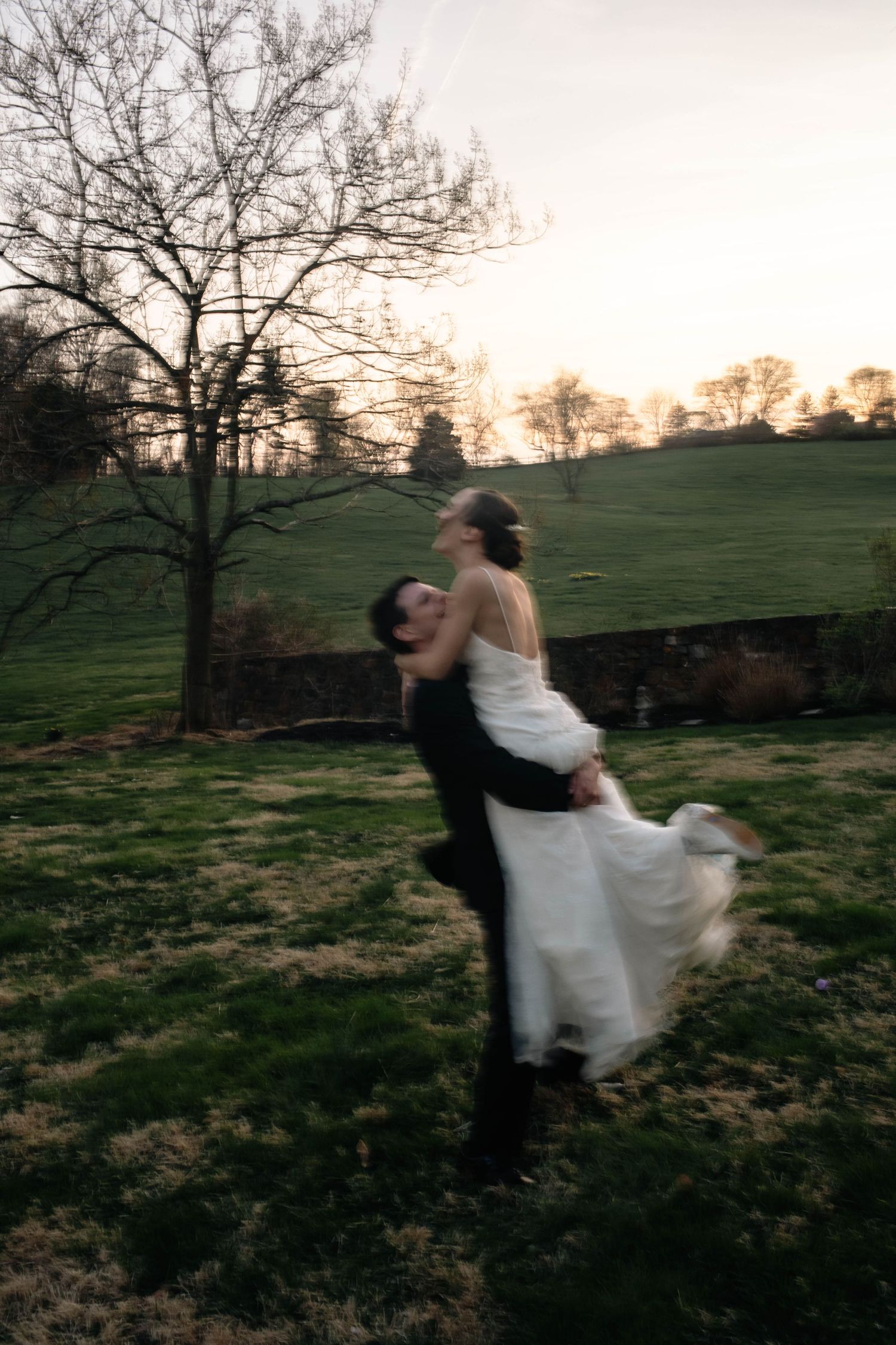 Groom joyfully lifts bride in white dress at sunset in a lush green outdoor wedding setting.
