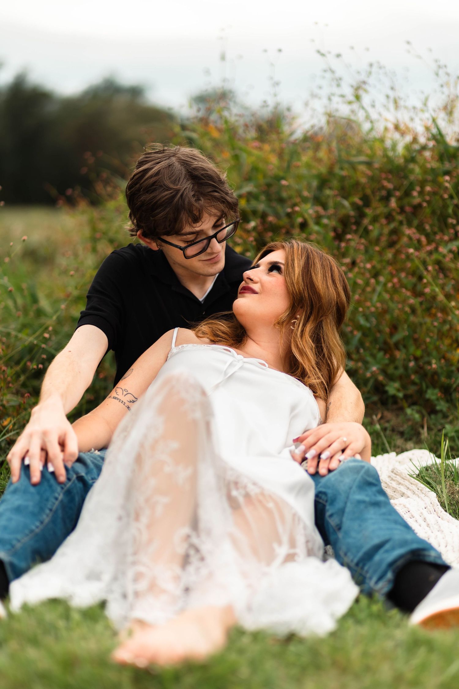 A couple relaxes together in a grassy field, woman in white lace dress leaning back against her partner during an engagement session in Oley