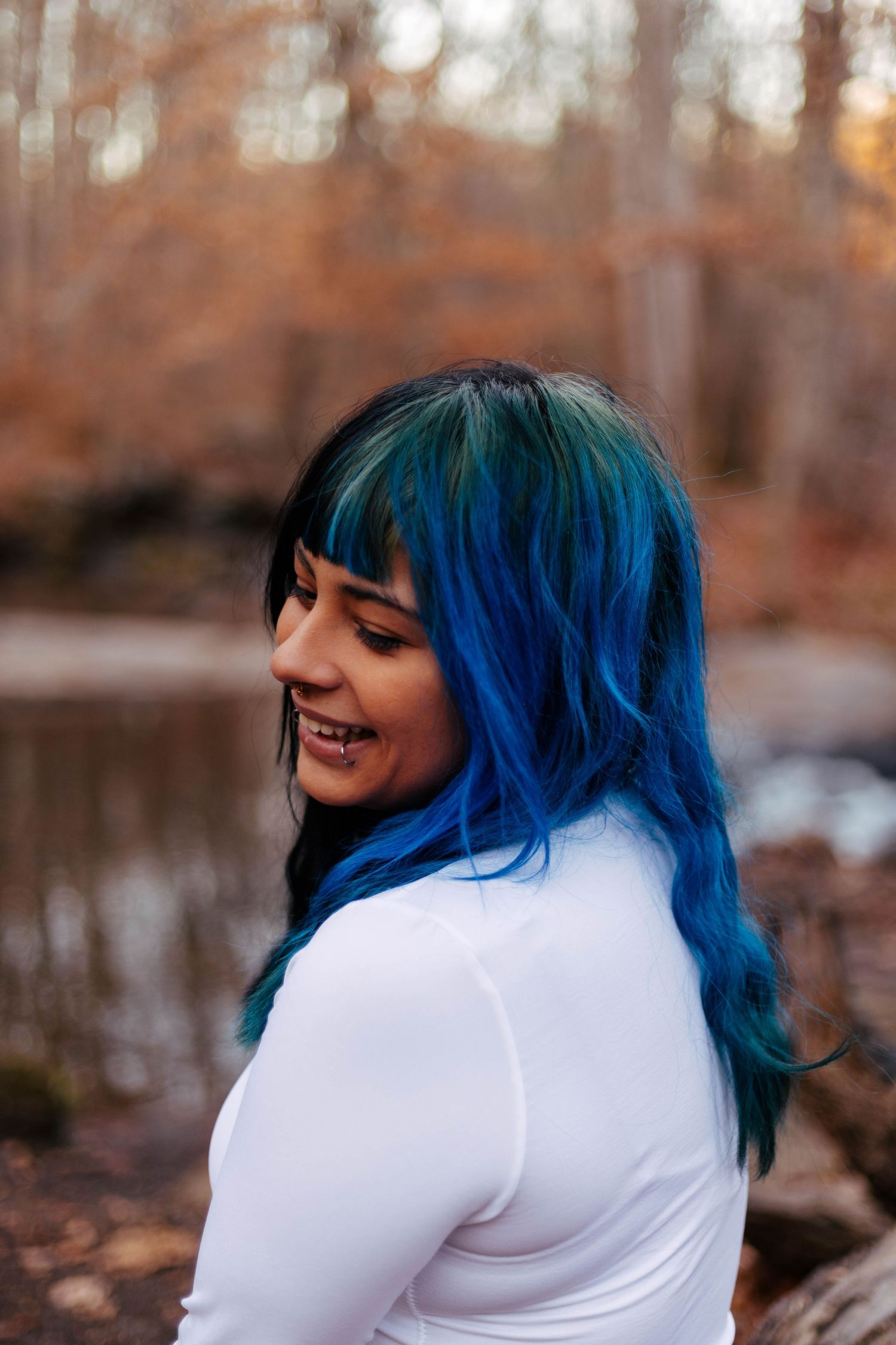 Young woman with vibrant blue and black ombre hair smiling outdoors in autumn forest setting.