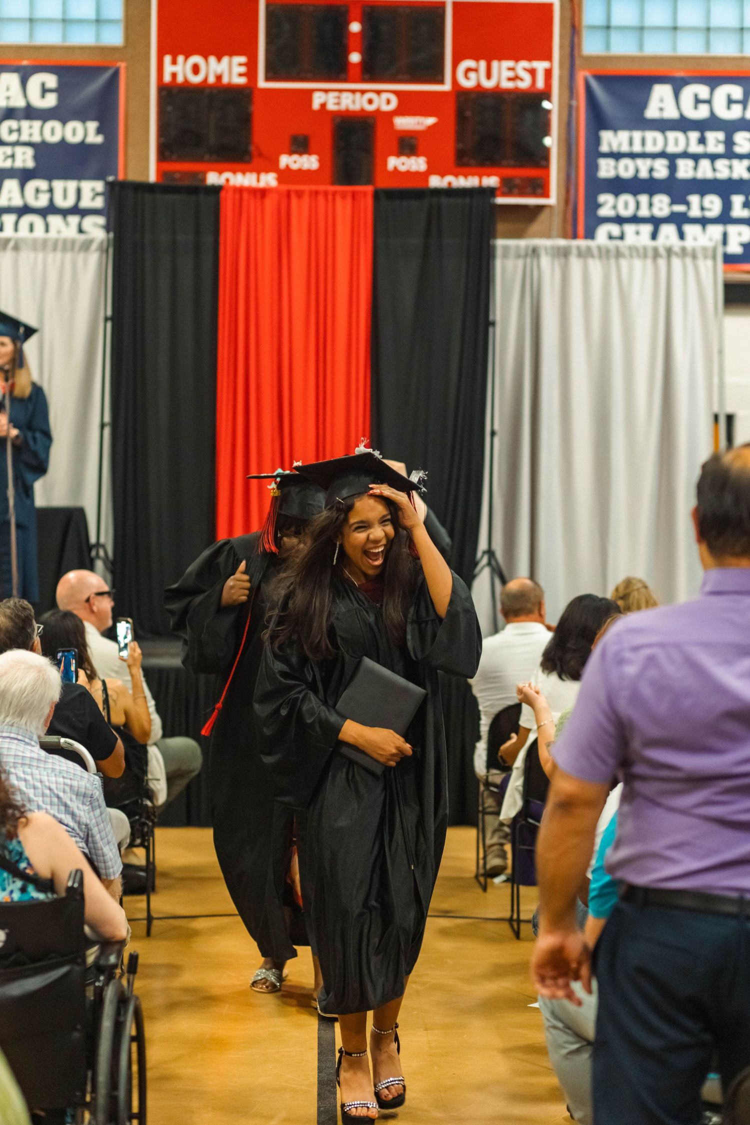 Smiling graduate in black cap and gown walking through aisle at graduation ceremony.