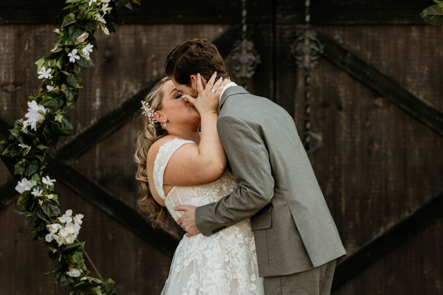 Bride and groom share their first kiss as husband and wife in front of a rustic wooden barn adorned with floral arch.