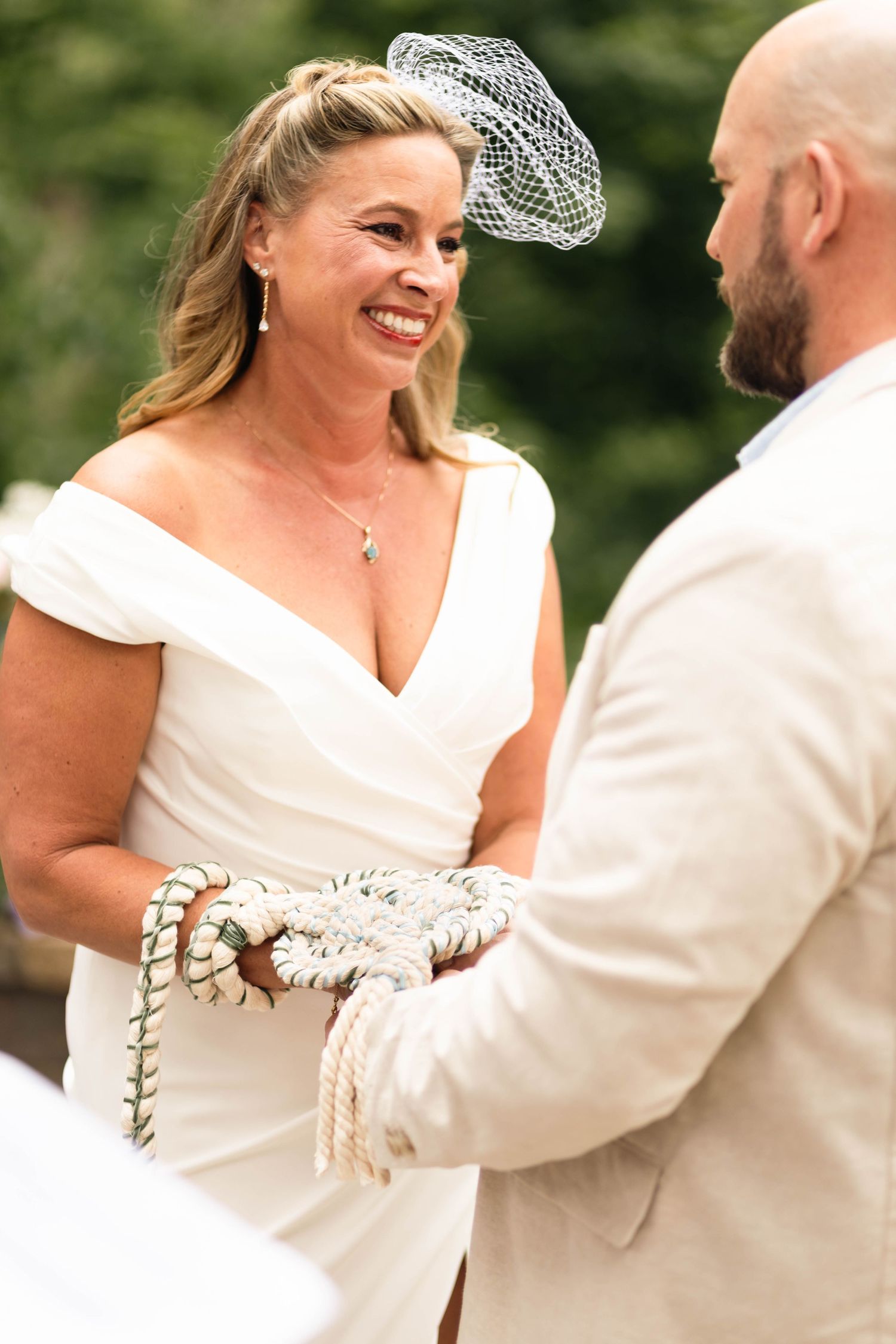 Smiling bride in white off-shoulder gown with birdcage veil during a handfasting wedding ceremony.