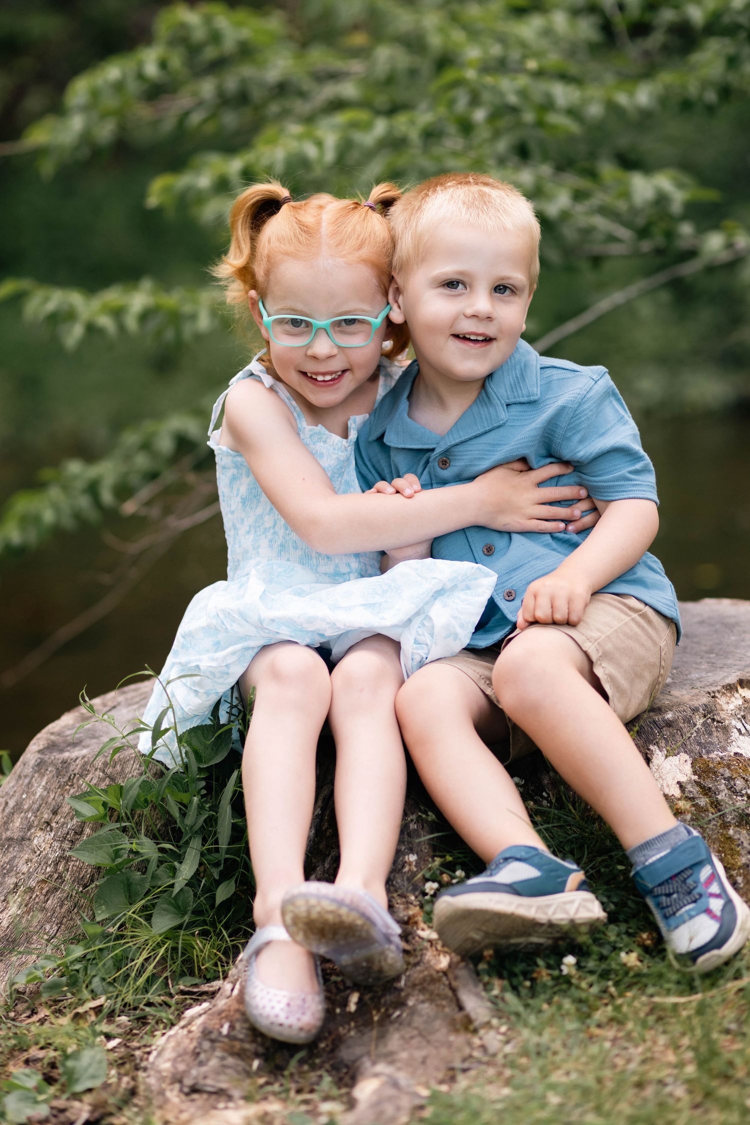 Red-haired girl with glasses hugs younger blond boy while sitting on rock in lush outdoor portrait in Boyertown, PA.