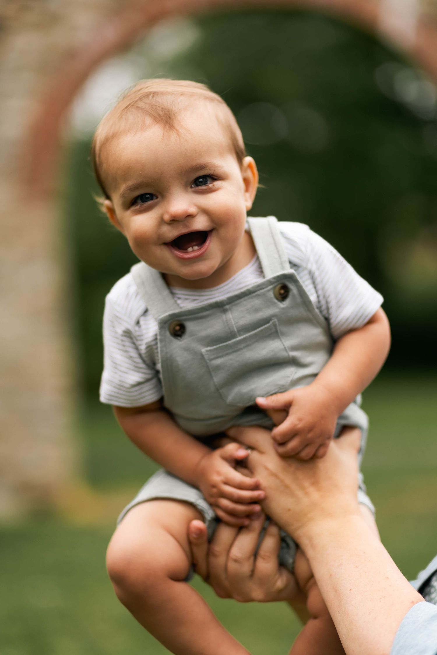 Smiling baby in gray overalls being lifted up by hands in an outdoor garden setting at Lockridge Park.