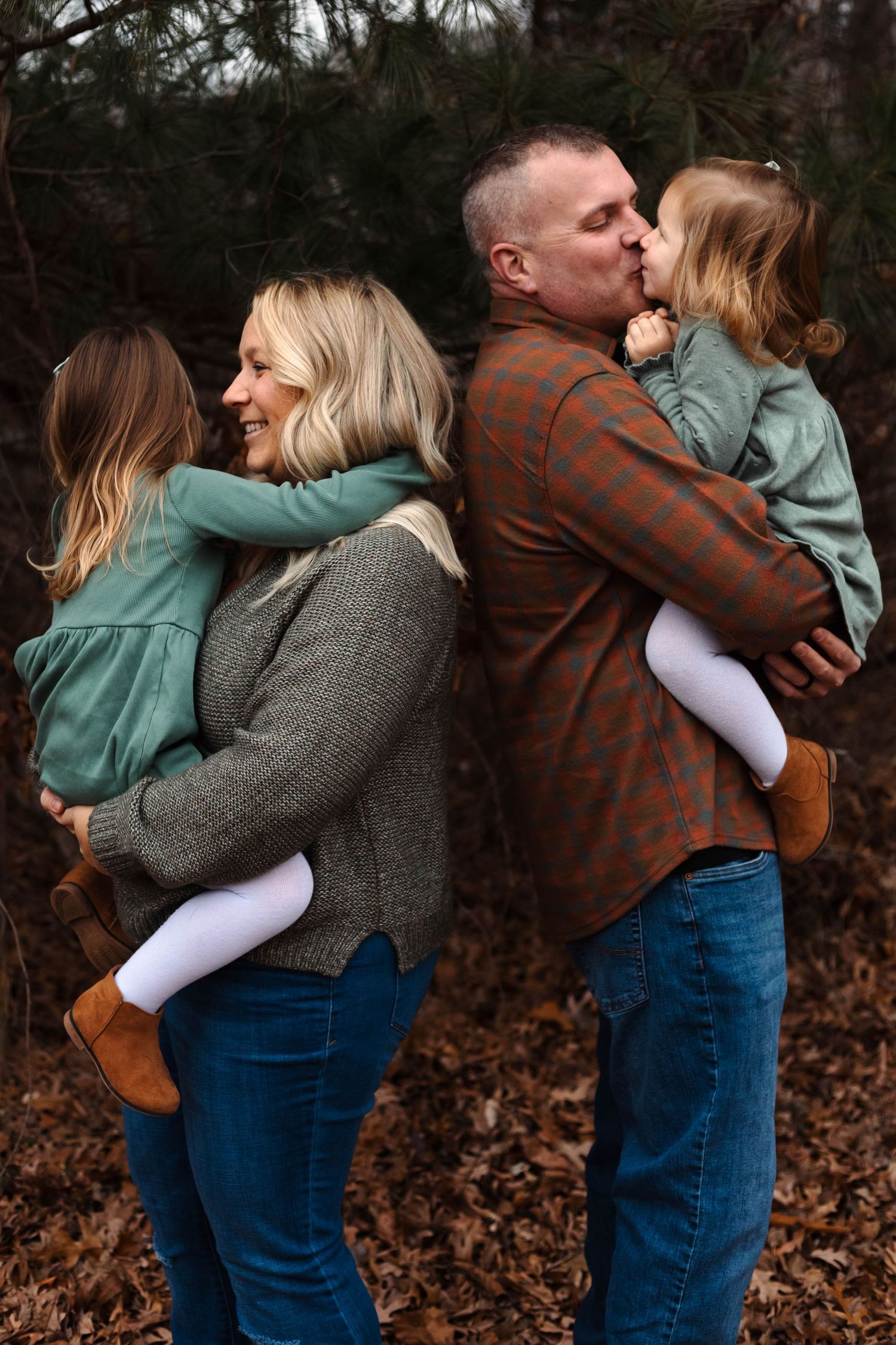 Parents each hold one of their two young daughters, sharing a playful back-to-back moment in an autumn forest at Green Lane Park.