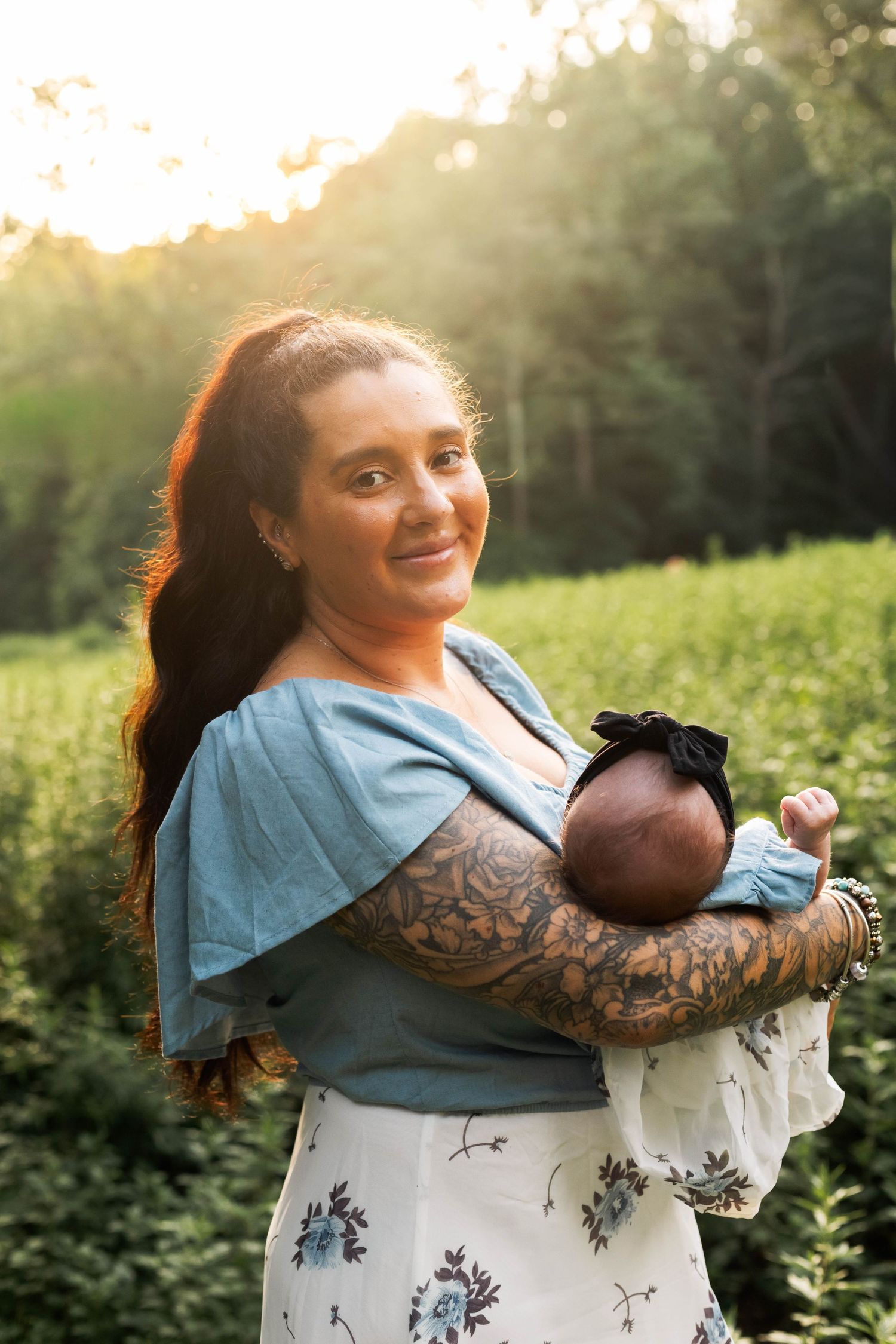 Smiling tattooed mother holding newborn baby wrapped in white blanket in golden outdoor light during a newborn session in Boyertown, PA.