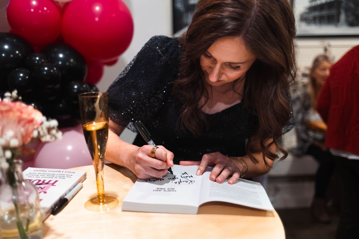 Brunette woman in black sequin dress signing a book at an event with balloons.