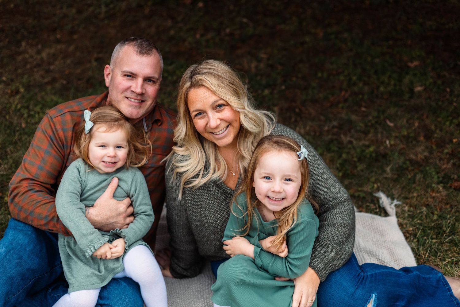 A happy family of four sits together on grass, with two young girls in matching green outfits smiling brightly at Green Lane Park.