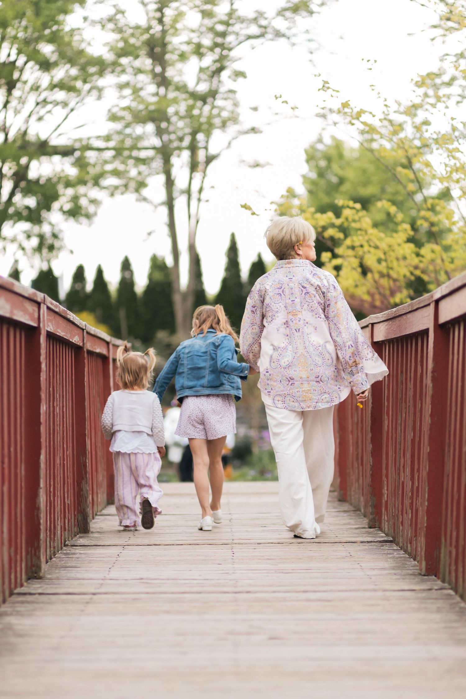 A grandmother walks hand-in-hand with two young girls across a charming red wooden bridge in a lush park at the Reading Museum Arboretum.