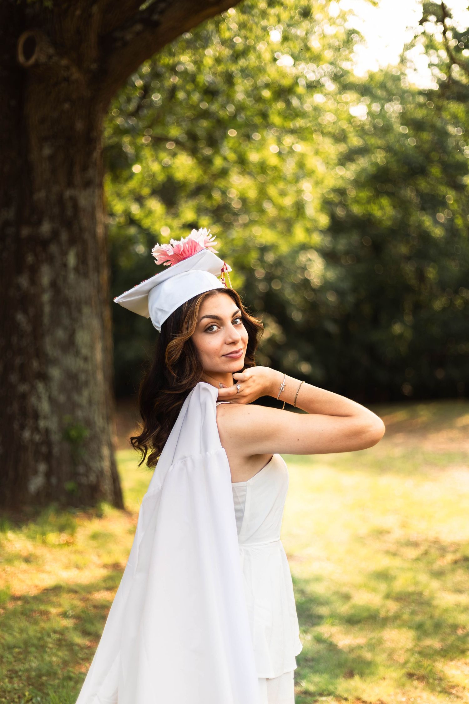 Young woman in white graduation cap and gown poses outdoors under green trees in a sunny park setting.