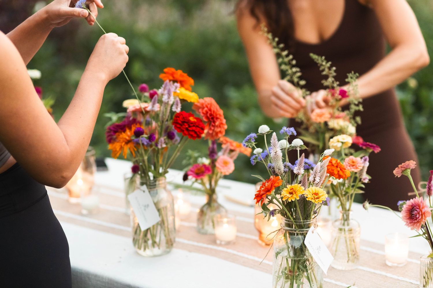 Women arrange vibrant wildflower bouquets in glass vases on a candlelit outdoor table.