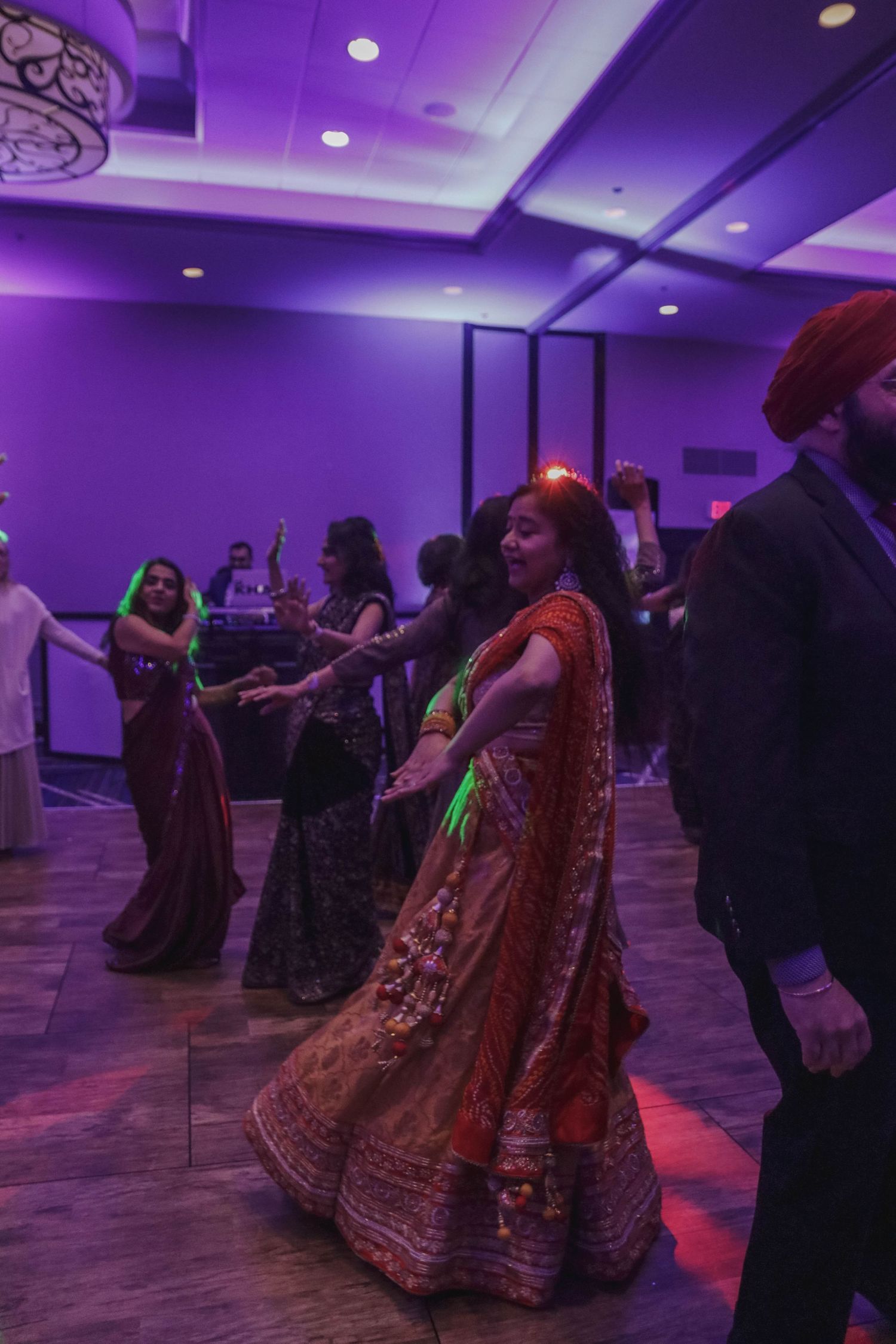 Woman in orange and red lehenga dancing with guests at a vibrant Indian wedding reception.