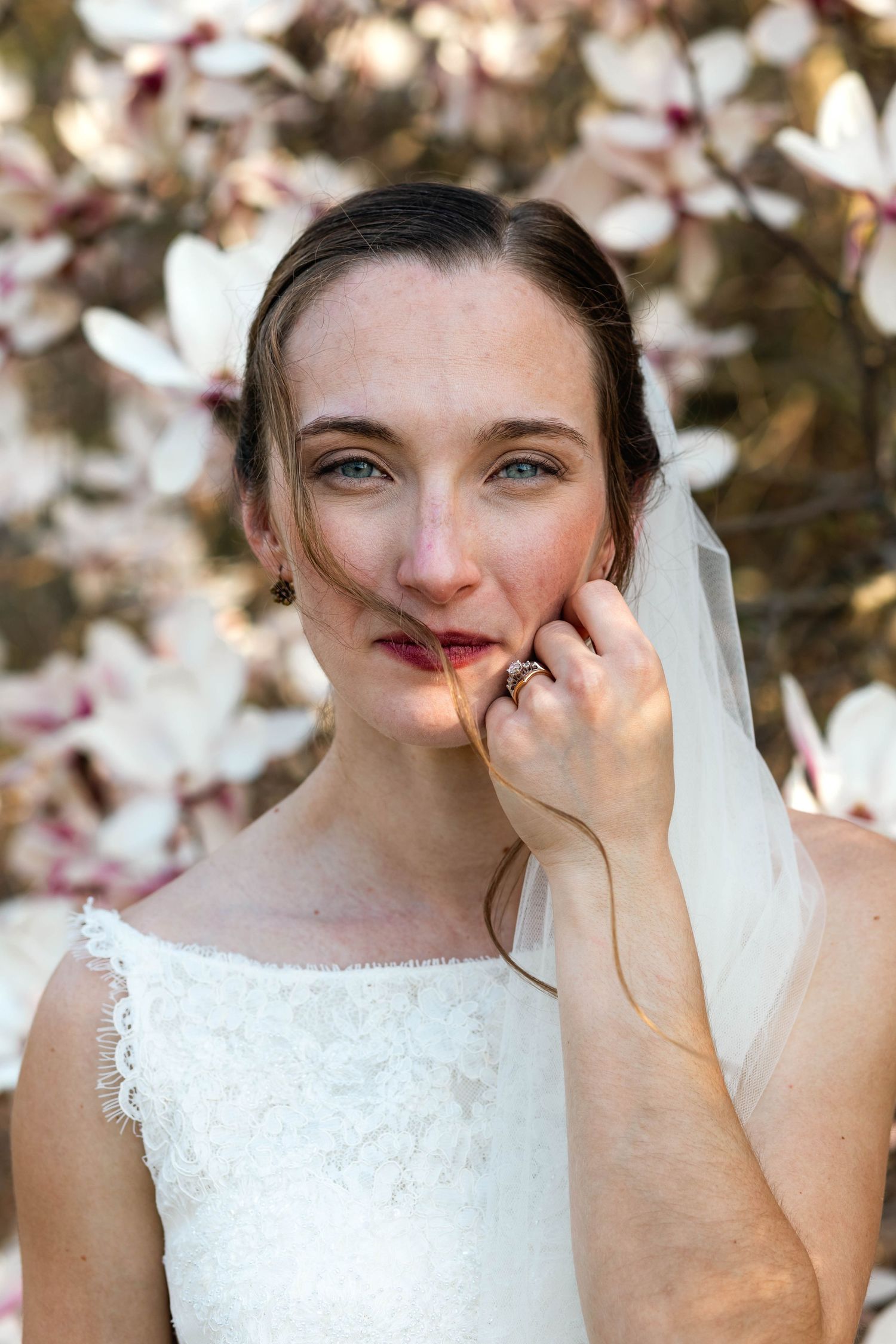 Beautiful blue-eyed bride in lace gown poses with veil near blooming magnolia flowers outdoors.