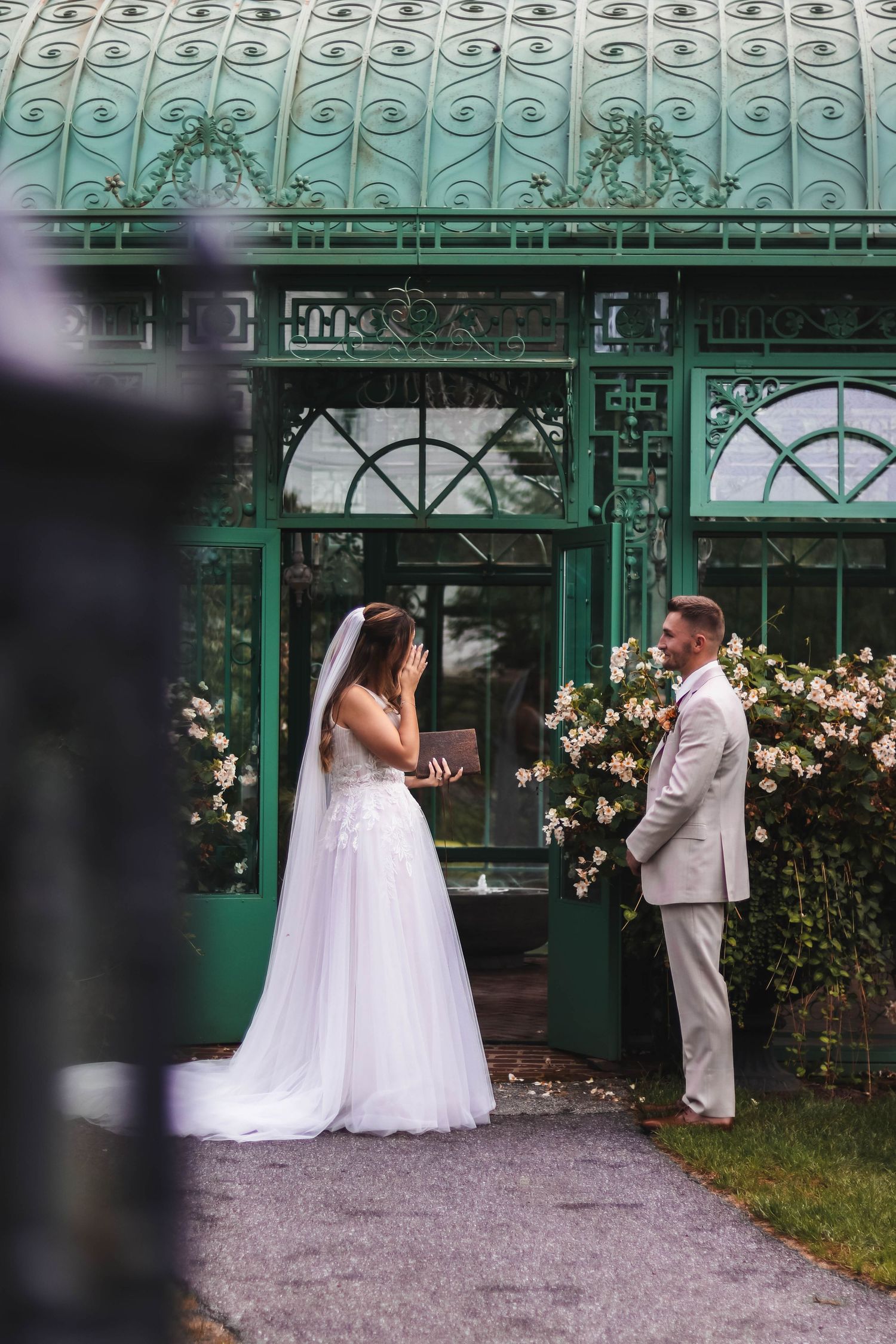 Bride reads vows from a book while groom listens in front of an ornate green greenhouse entrance in Lancaster.