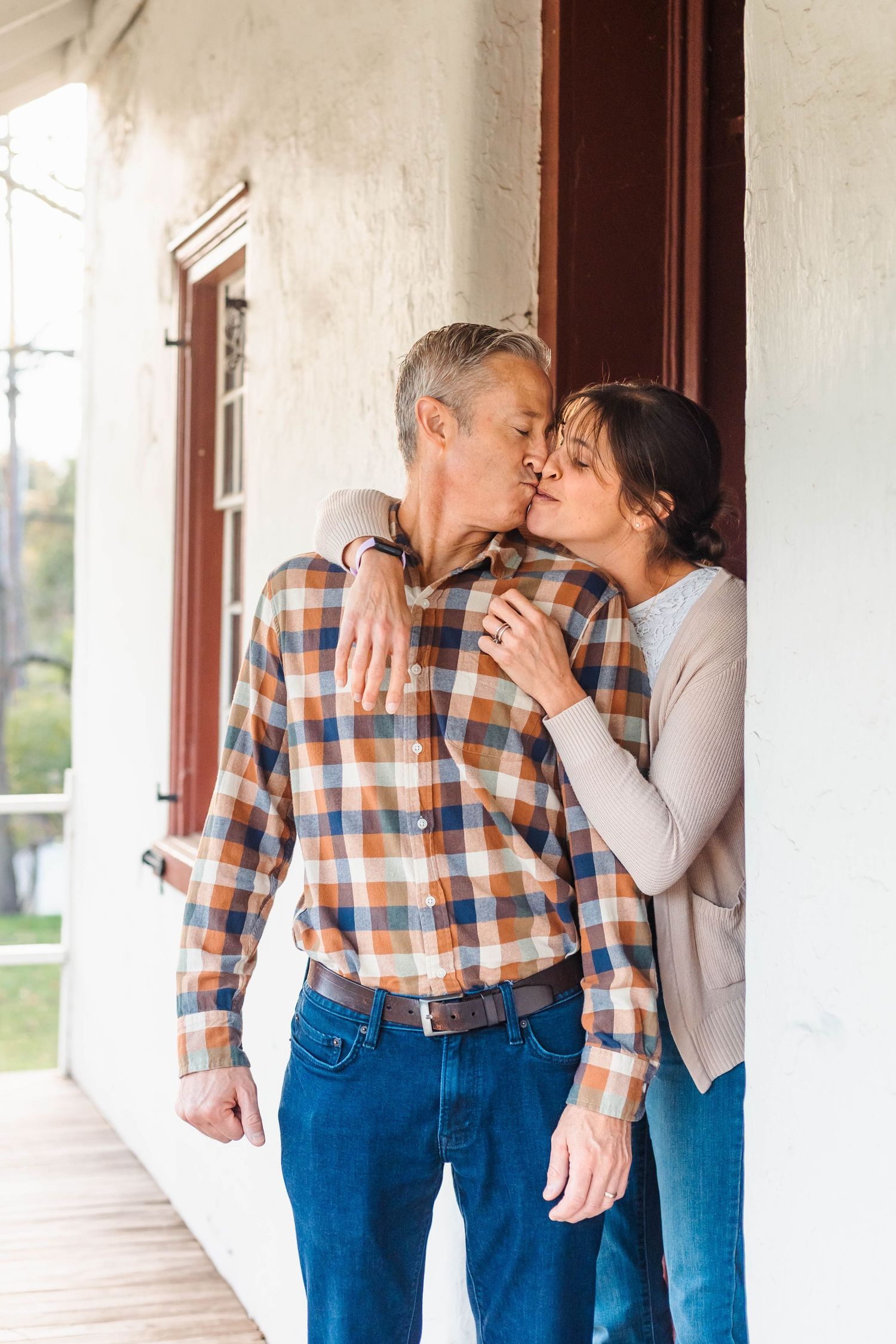 A couple shares a kiss while leaning against a white building wall on a porch in Harleysville, PA.