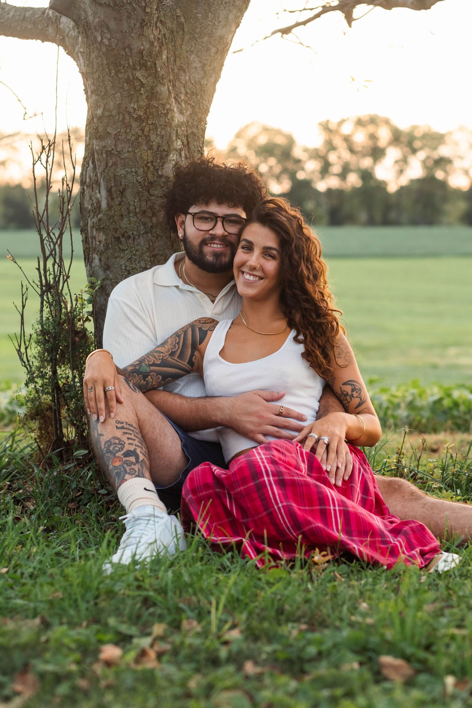 Happy couple sitting together under a tree at sunset, man with tattoos embracing smiling woman during an engagement session in Elverson, PA.