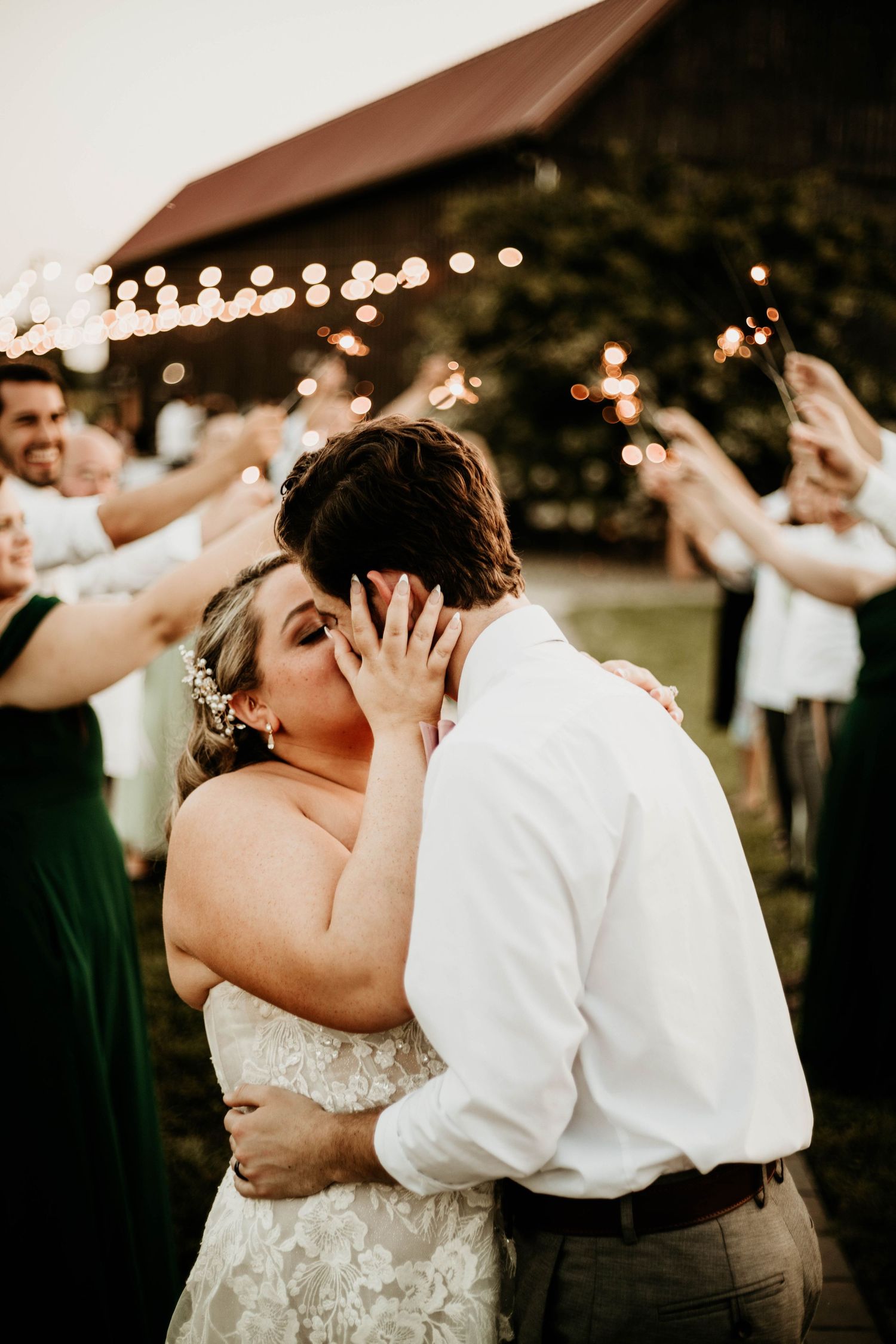 Newlyweds sharing first kiss surrounded by sparkler-holding guests outside rustic barn.