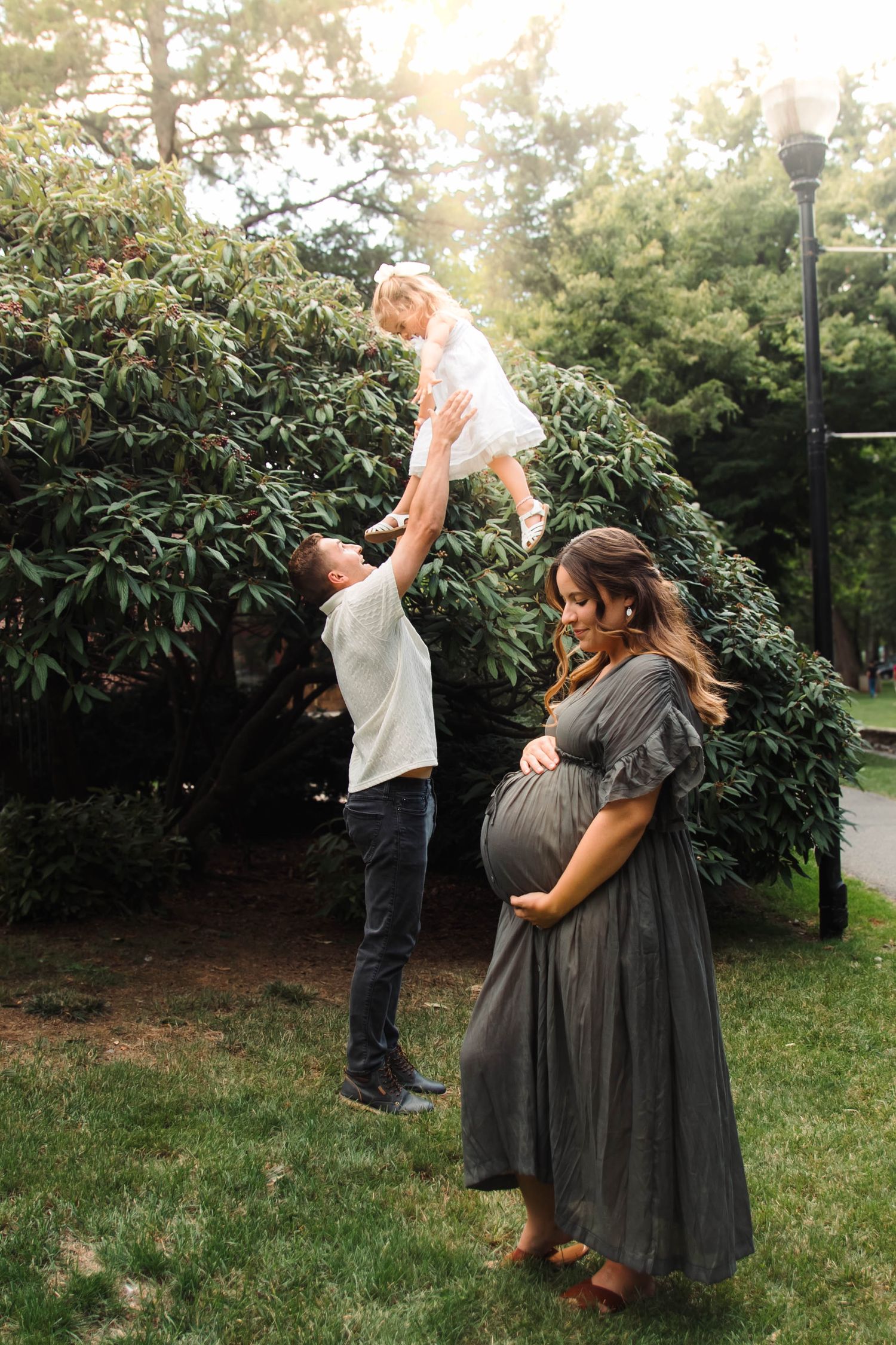 Father lifts toddler in white dress while pregnant mother in gray gown cradles bump in sunny park.