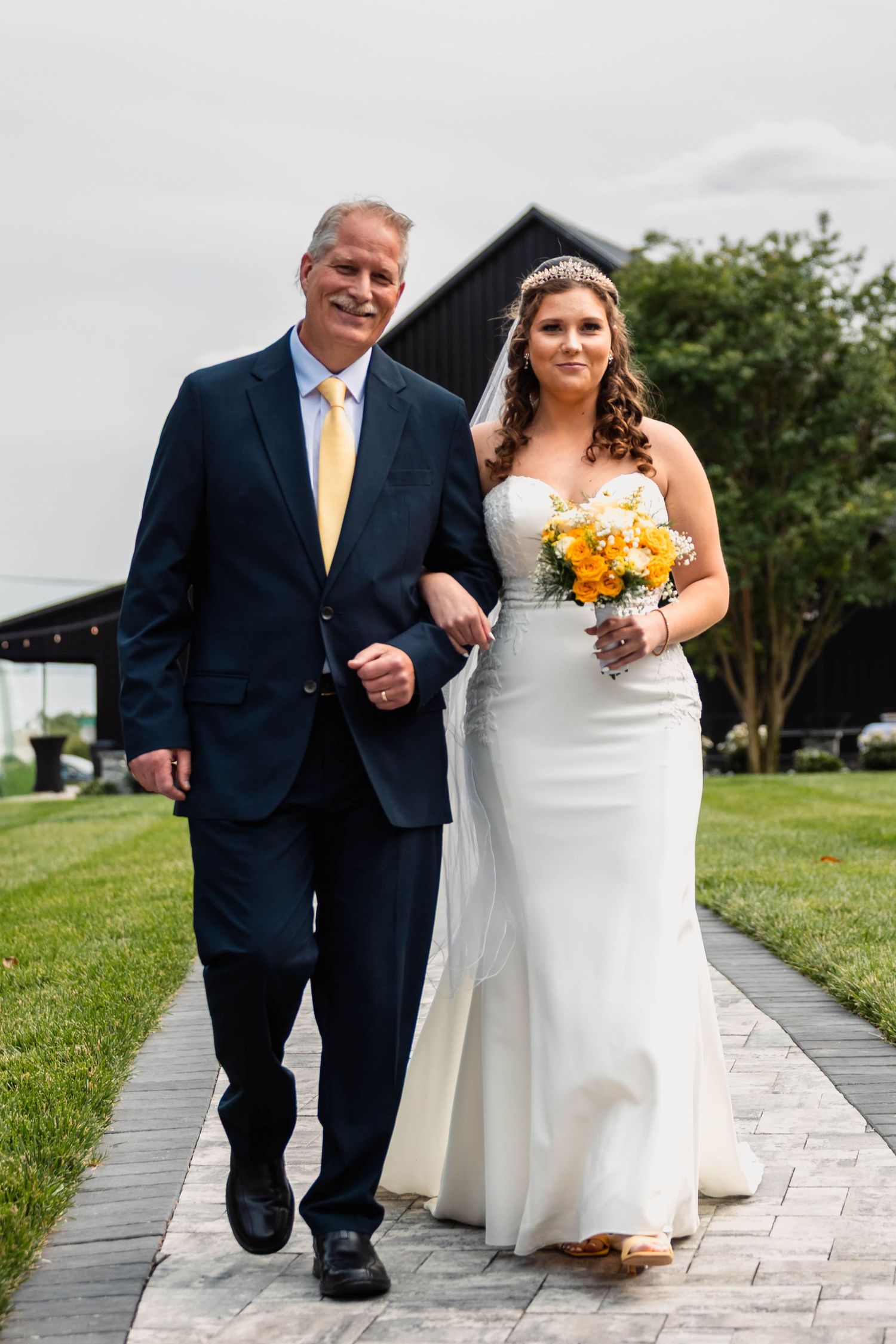 Bride in white gown holding yellow bouquet walks down aisle with father in navy suit and yellow tie.