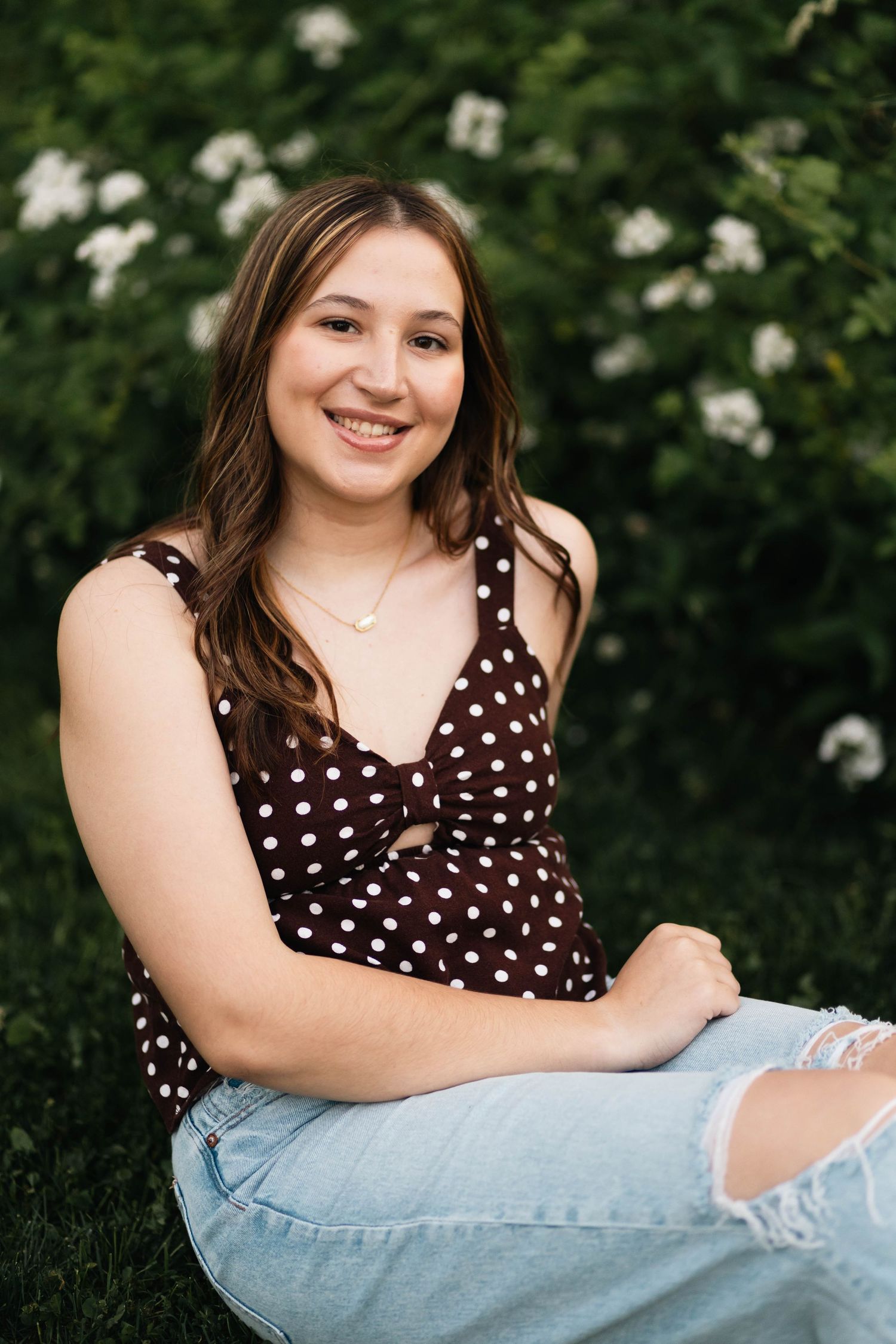 Boyertown High School senior in brownpolka dot top and ripped jeans sitting outdoors near white flowering bushes.