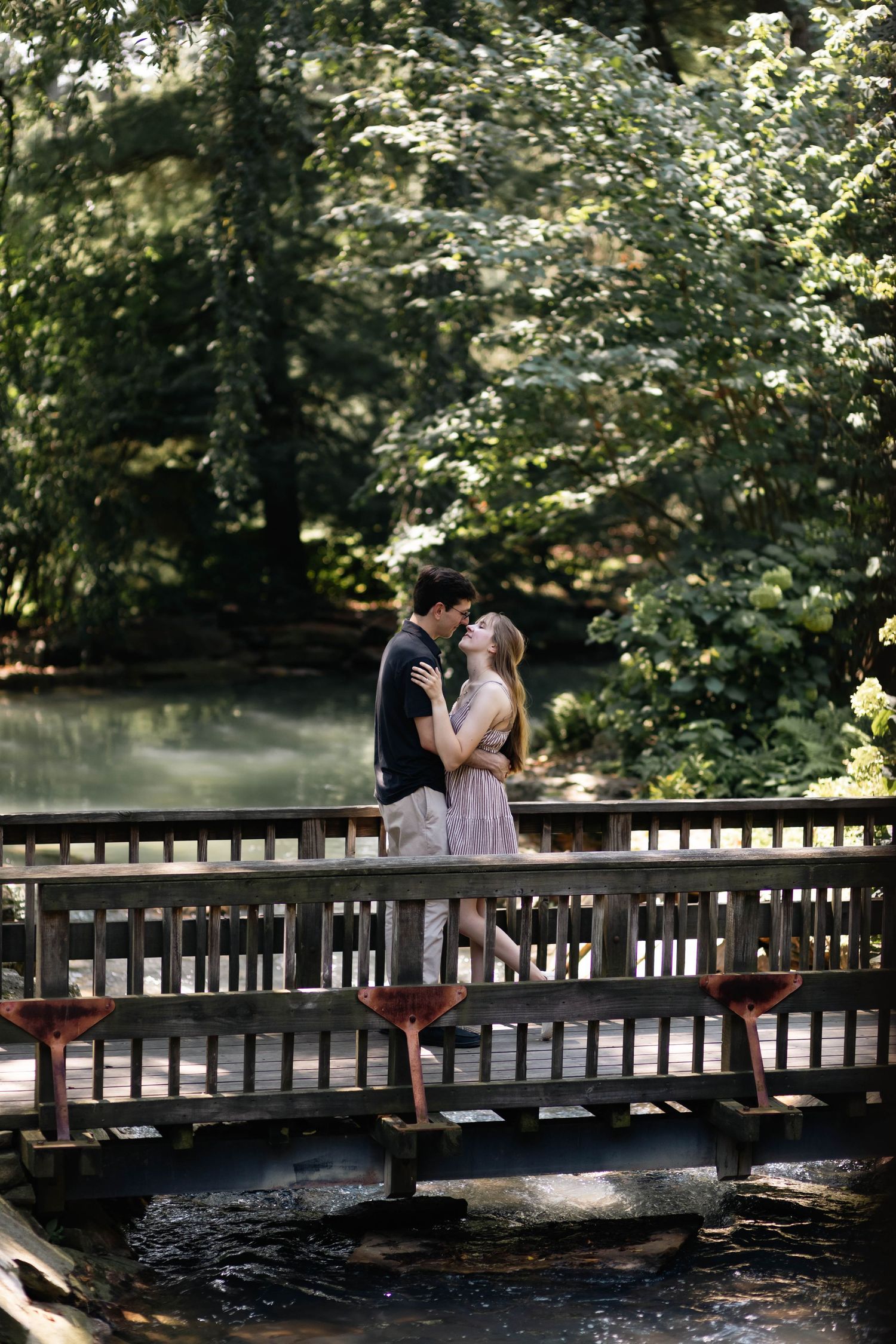 Couple sharing a romantic kiss on a wooden bridge surrounded by lush green trees and a serene stream.