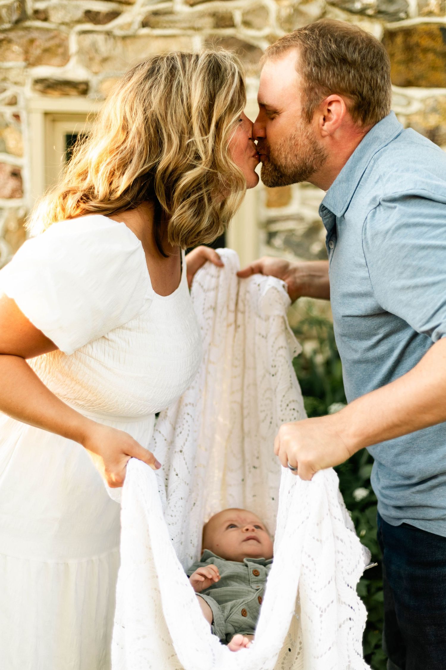 Loving parents kissing over their newborn baby wrapped in a white knit blanket outdoors.