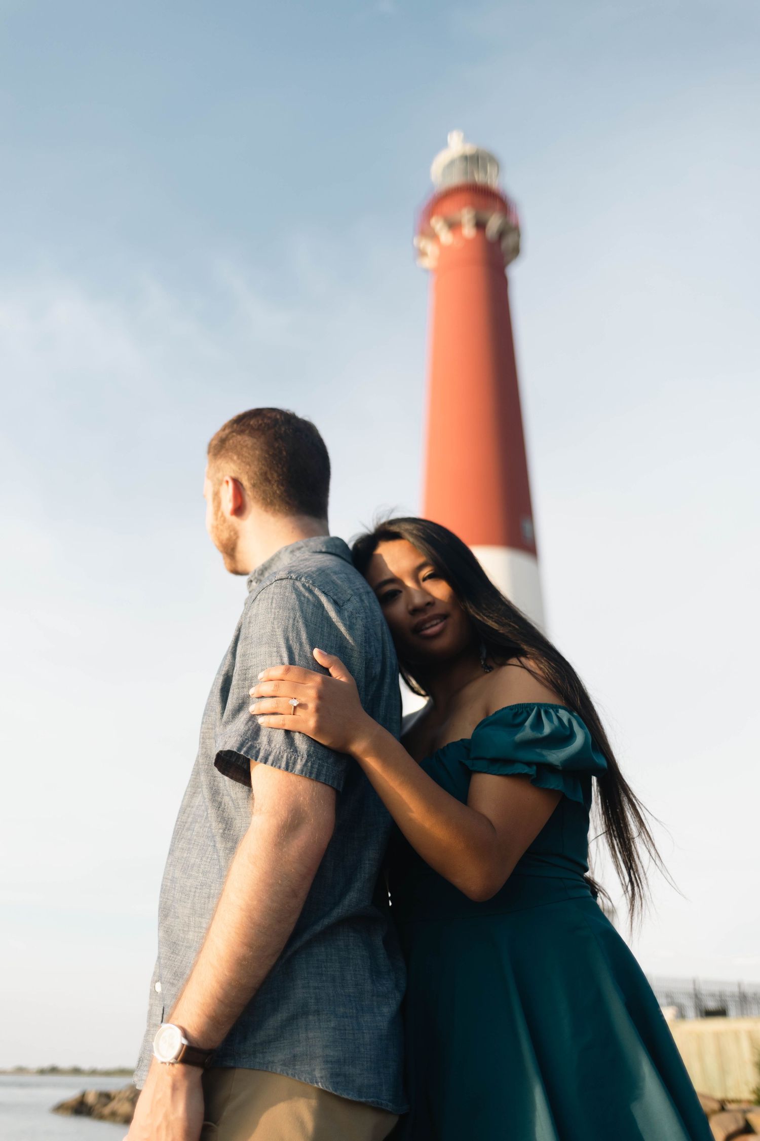 Woman in teal dress embraces man from behind near a tall red and white lighthouse in Barnegat, NJ.