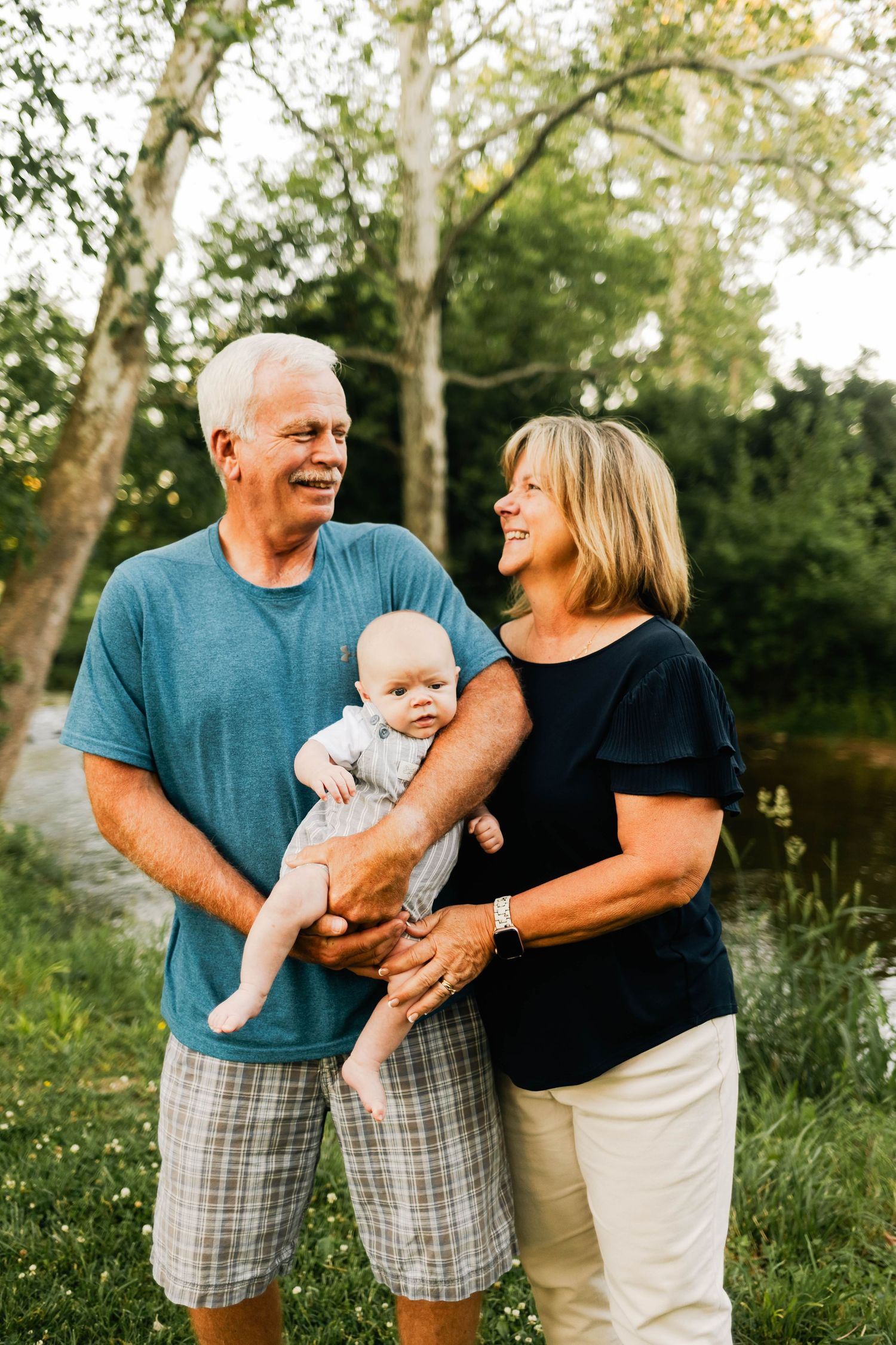 Happy grandparents smiling at each other while holding their infant grandchild outdoors near trees in Elverson, PA.