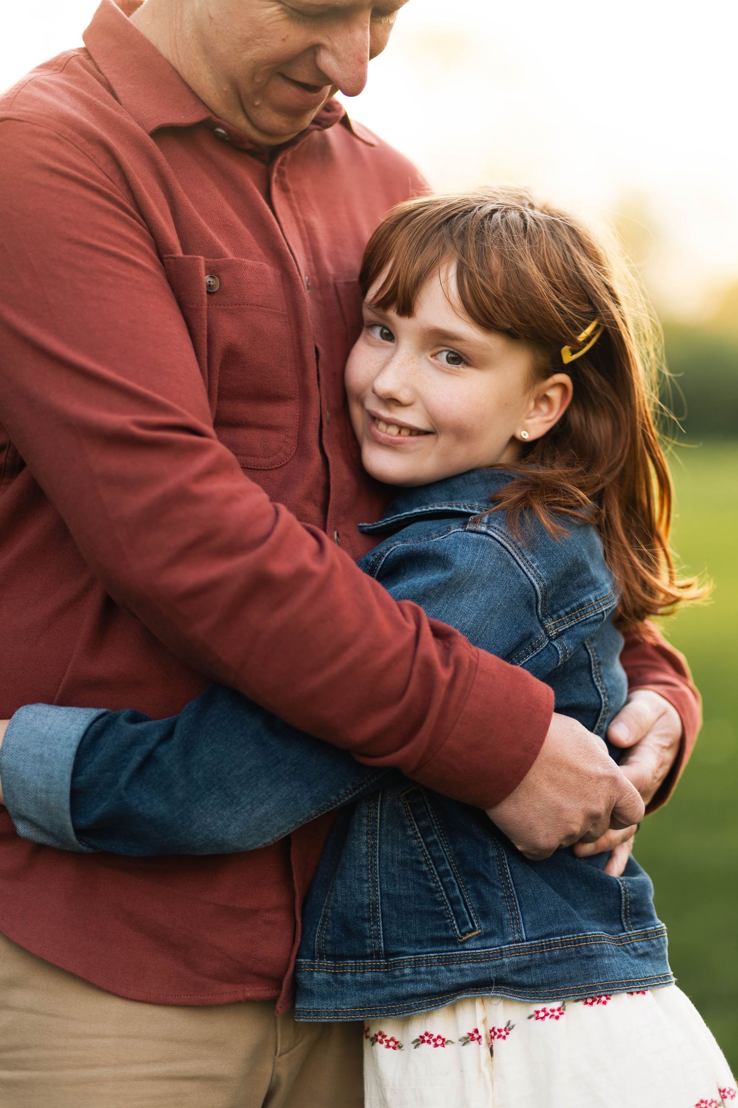 A young red-haired girl in a denim jacket smiles while being hugged by a man in a red shirt outdoors in Elverson, PA.