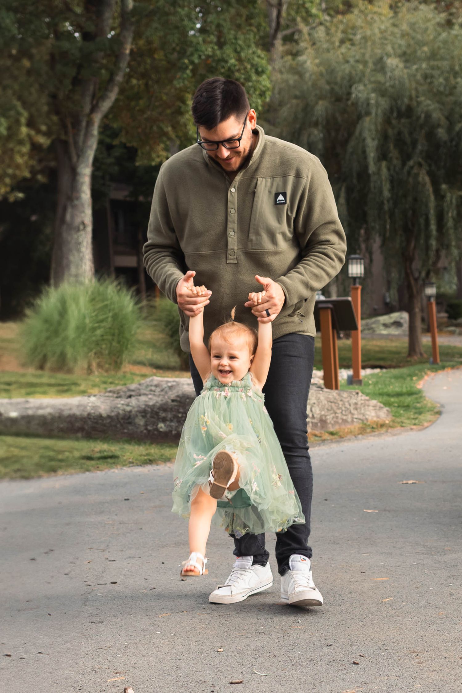 A father helps his toddler daughter take steps on a park path, both smiling in an outdoor setting in the Poconos.