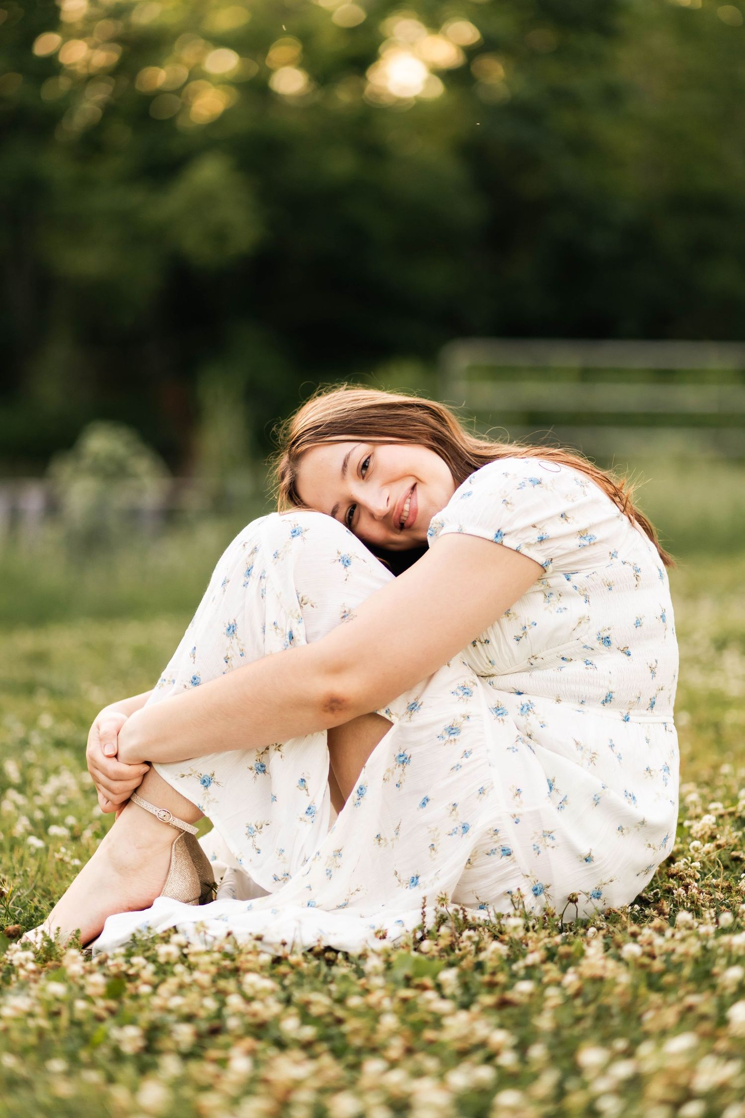 Cheerful young woman in floral white dress sitting in a clover field, hugging her knees and smiling.