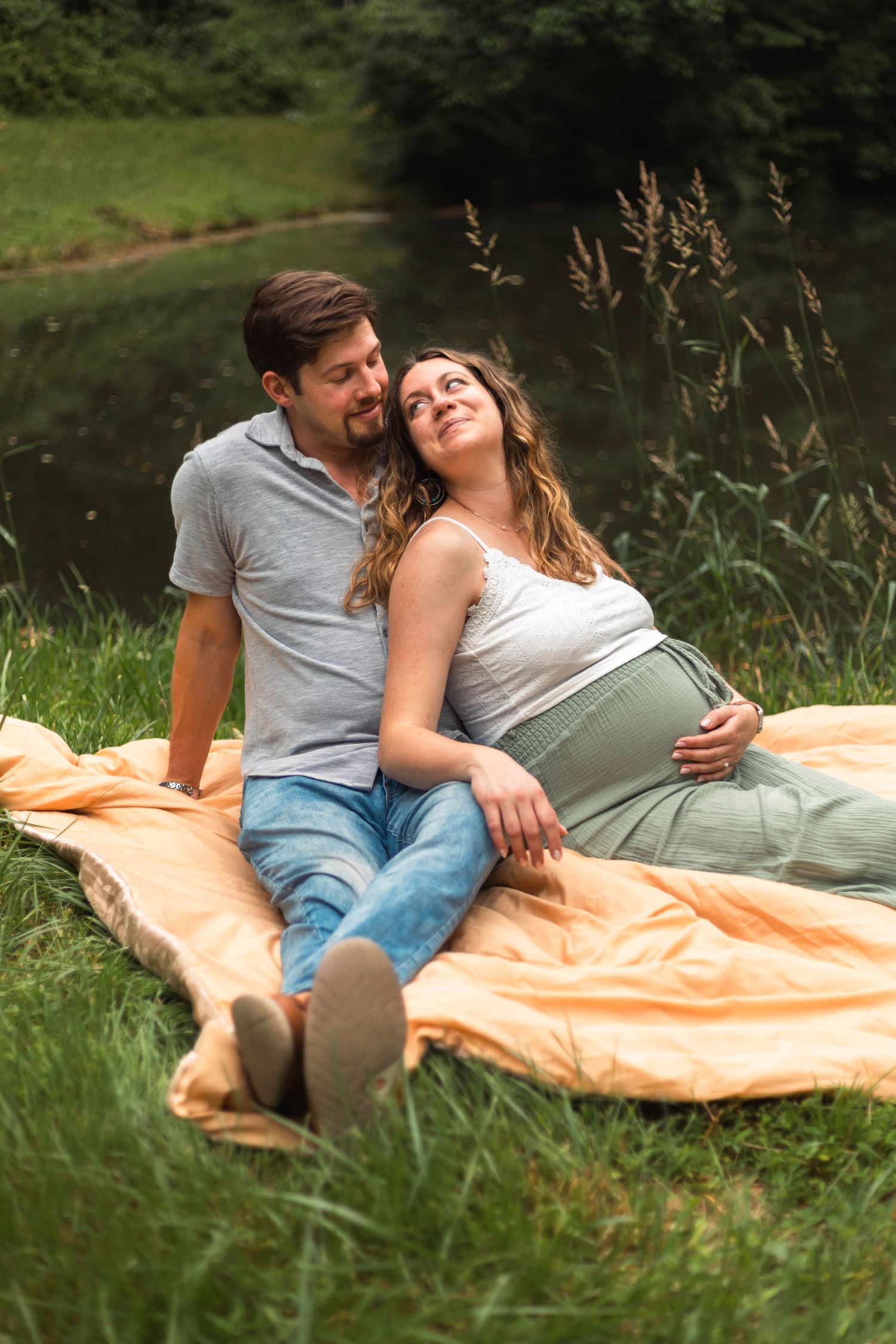 Happy pregnant couple relaxing on orange blanket by a pond surrounded by lush green grass.